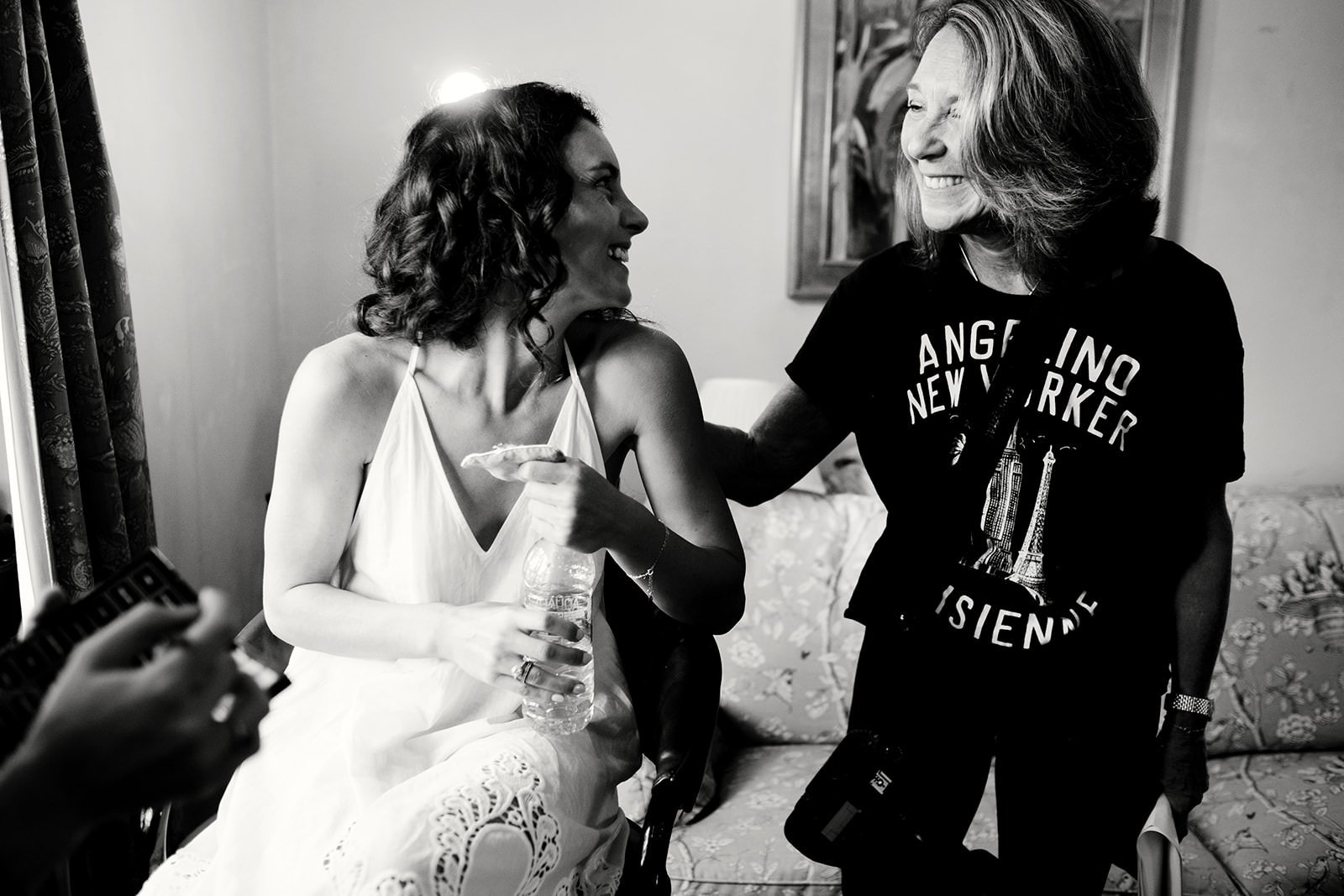 Two women with curly hair, smiling and engaging in conversation inside a room, one seated holding a water bottle, and the other standing wearing a black t-shirt with city skyline and landmark illustrations.