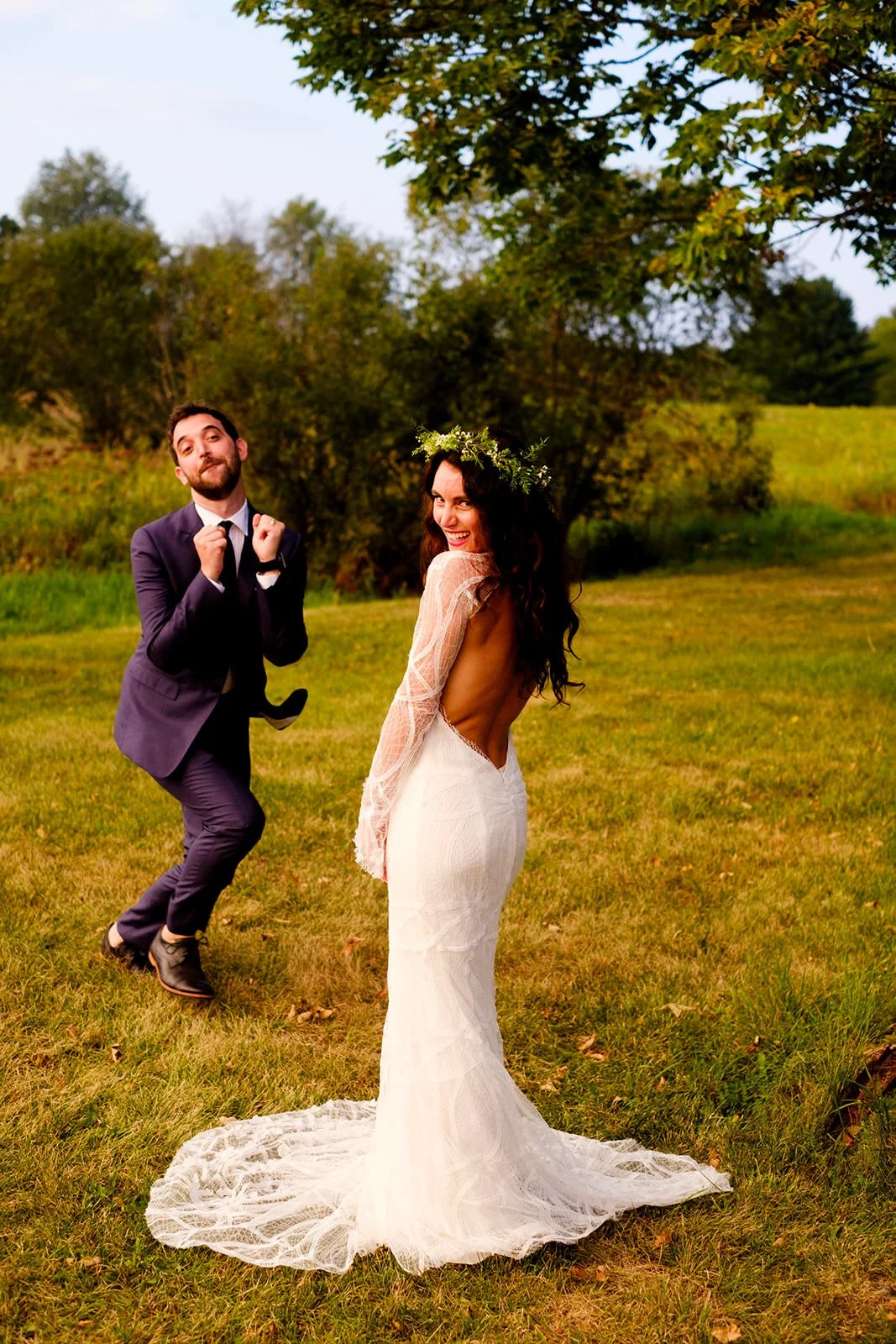 A joyful bride in a white dress with an open back and lace sleeves is posing outdoors on a grassy field, wearing a green floral crown. A playful man in a dark suit and tie is dancing behind her, making a fun expression. Trees and green landscape are 