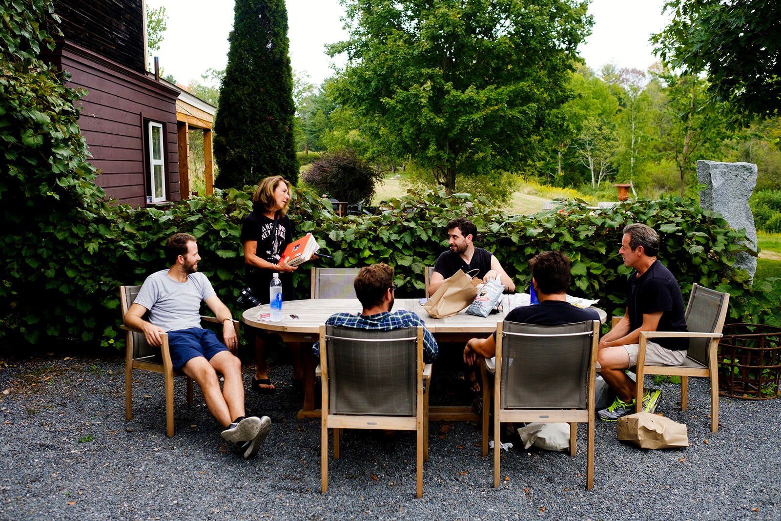 Group of six people sitting around a circular outdoor table in a garden, with a woman standing and speaking to them. The setting includes lush green trees and a house with a purple exterior in the background.