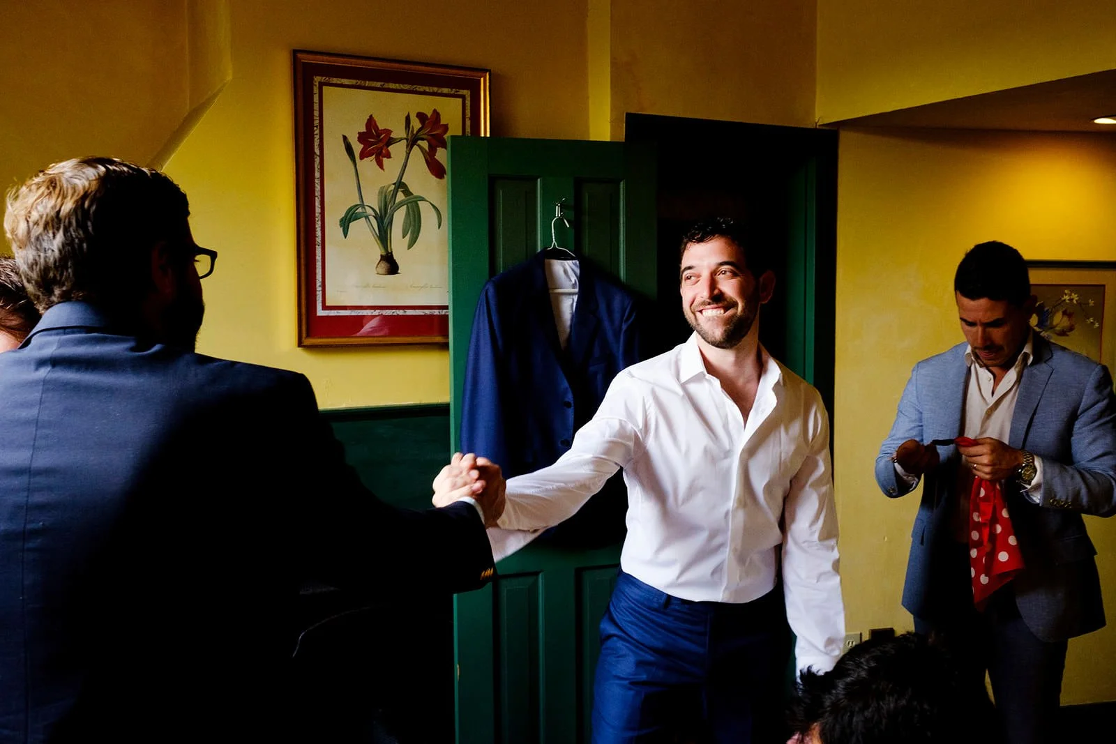 Man in white shirt shaking hands with man in blue suit in a room with yellow walls and framed flower artwork.
