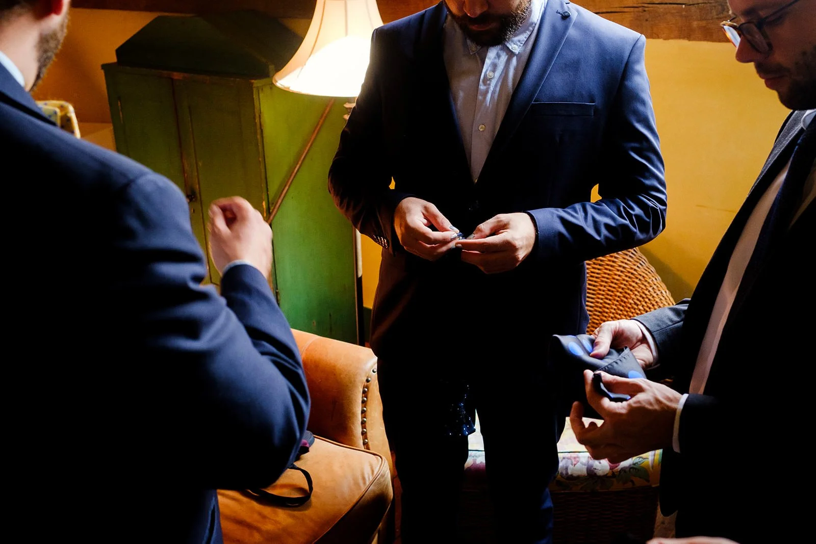 Three men in suits are gathered in a warmly lit room, focusing on their phones and a small object one man is holding.