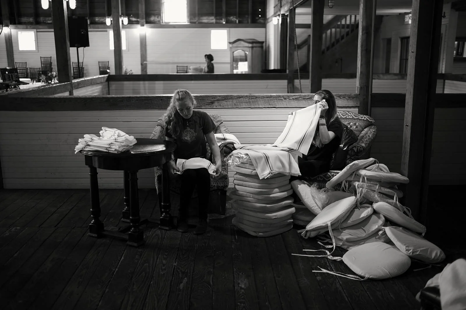 Two women are seated indoors among stacks of folded linens or garments. One woman is folding clothes on a small table, and the other is sitting on a cushioned chair, holding a fabric up to her face. The background shows a rustic interior with wooden 