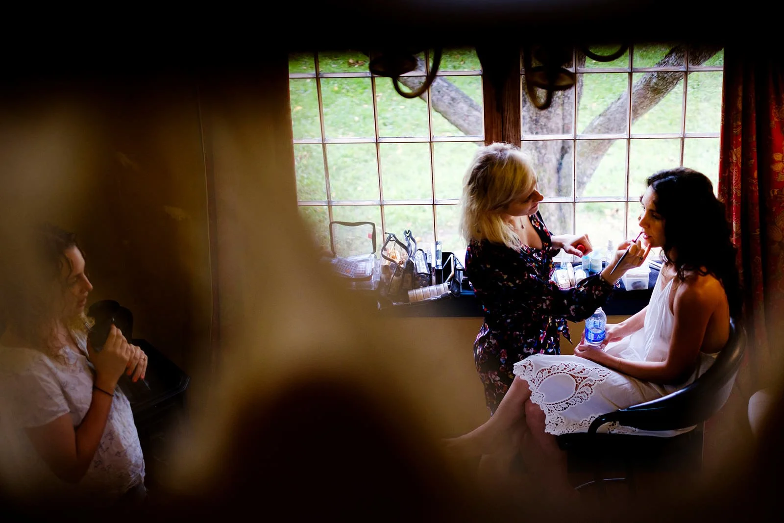 A makeup artist applies makeup on a woman sitting in a chair by a large window, with another girl holding a microphone nearby.