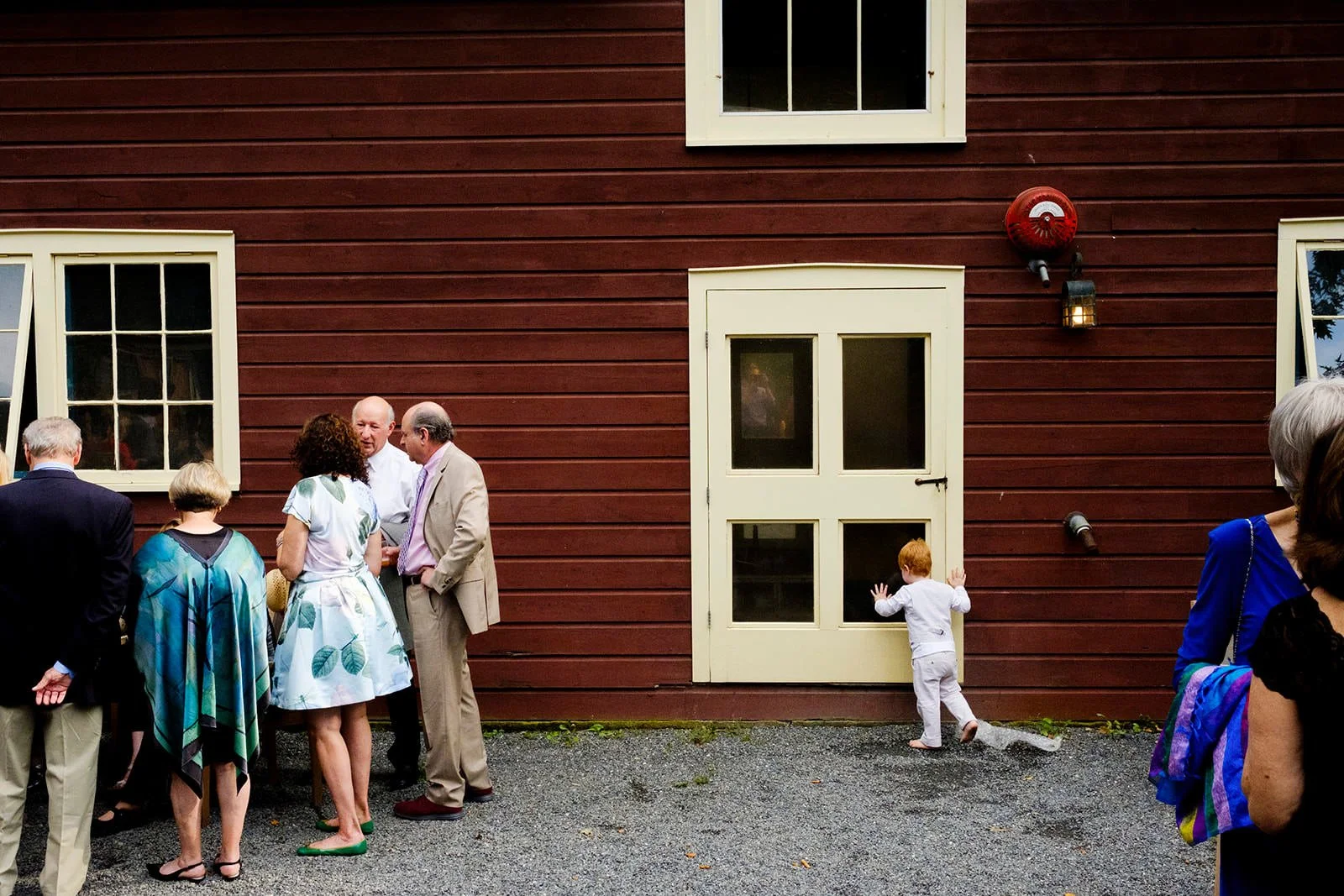 Group of people socializing outside a red wooden building with beige trim, including two children near the door, one of them with hands on the door, and several adults chatting.