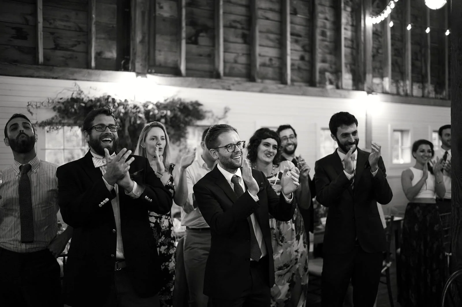 Group of people dressed in formal attire, clapping and smiling at an indoor event with string lights and wooden walls.
