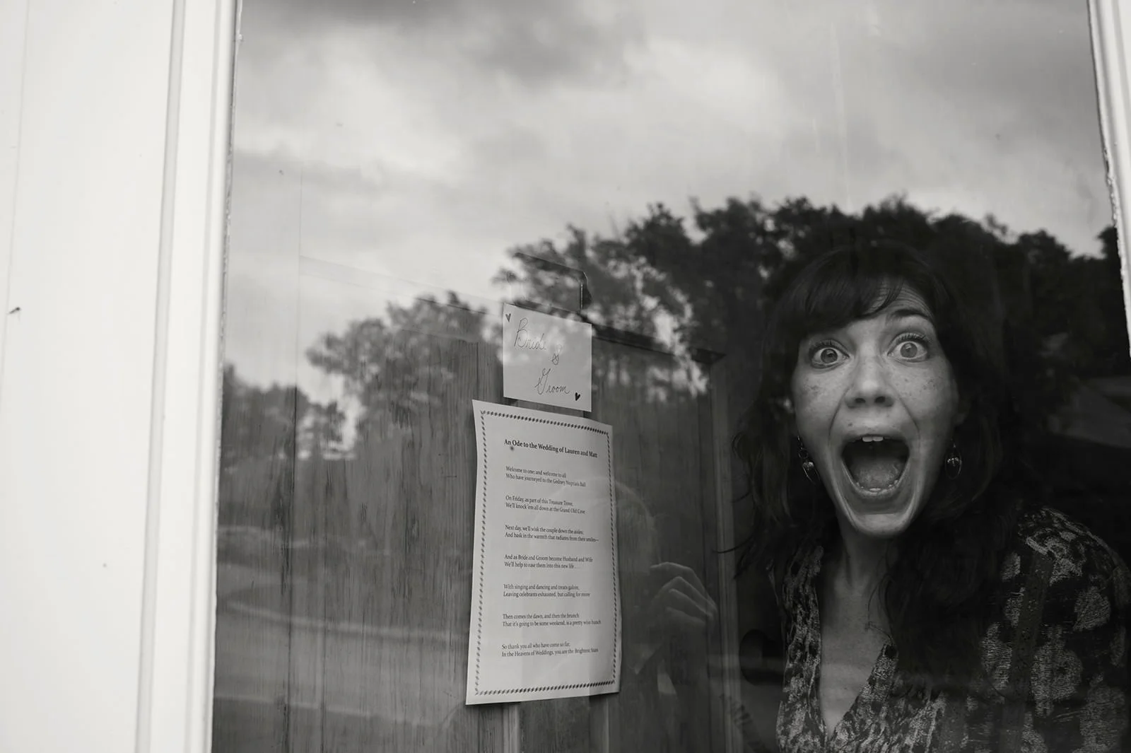 A woman with dark hair and earrings is seen behind a glass window, her mouth wide open in surprise or excitement. In the background, trees and a cloudy sky are reflected on the glass. There are also some papers and notes taped on the glass.