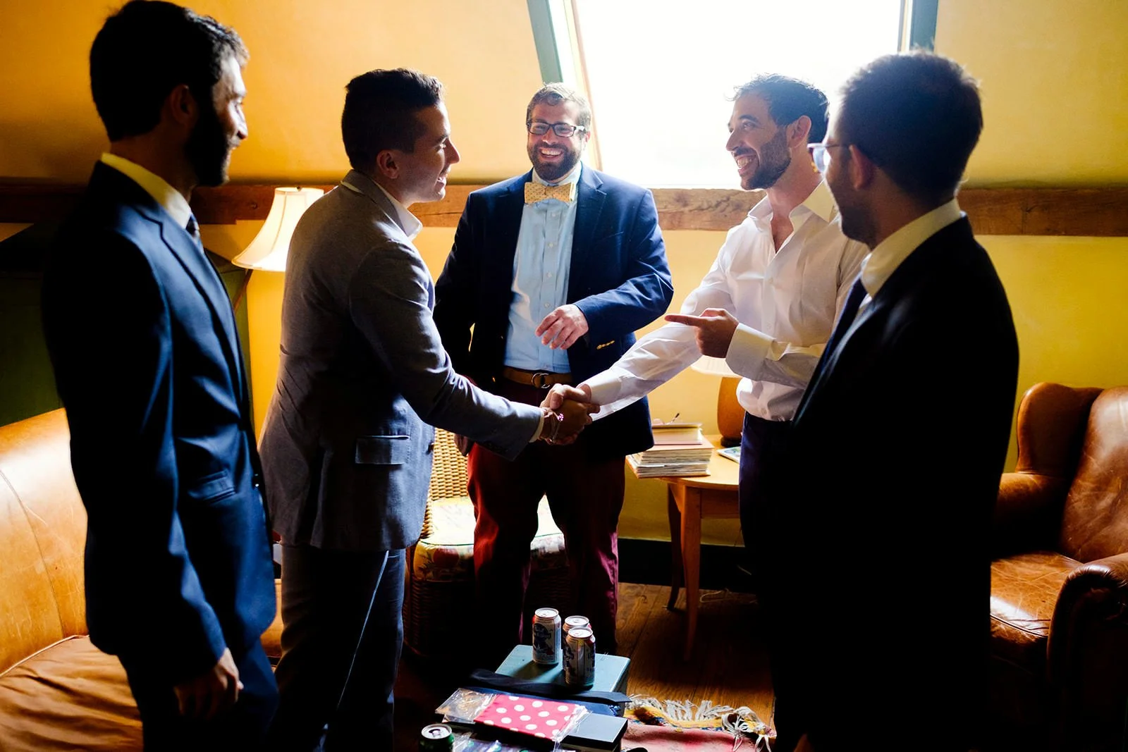 Six men dressed in suits, one wearing a boutonniere, standing in a room with yellow walls, shaking hands and smiling, with one man pointing.