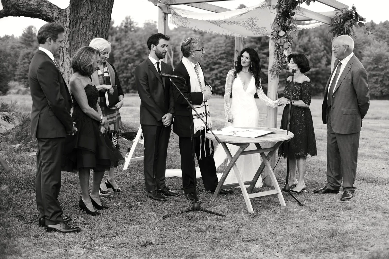 A black and white photo of a wedding ceremony outdoors with a wedding couple, officiant, and guests under a decorated wooden arch