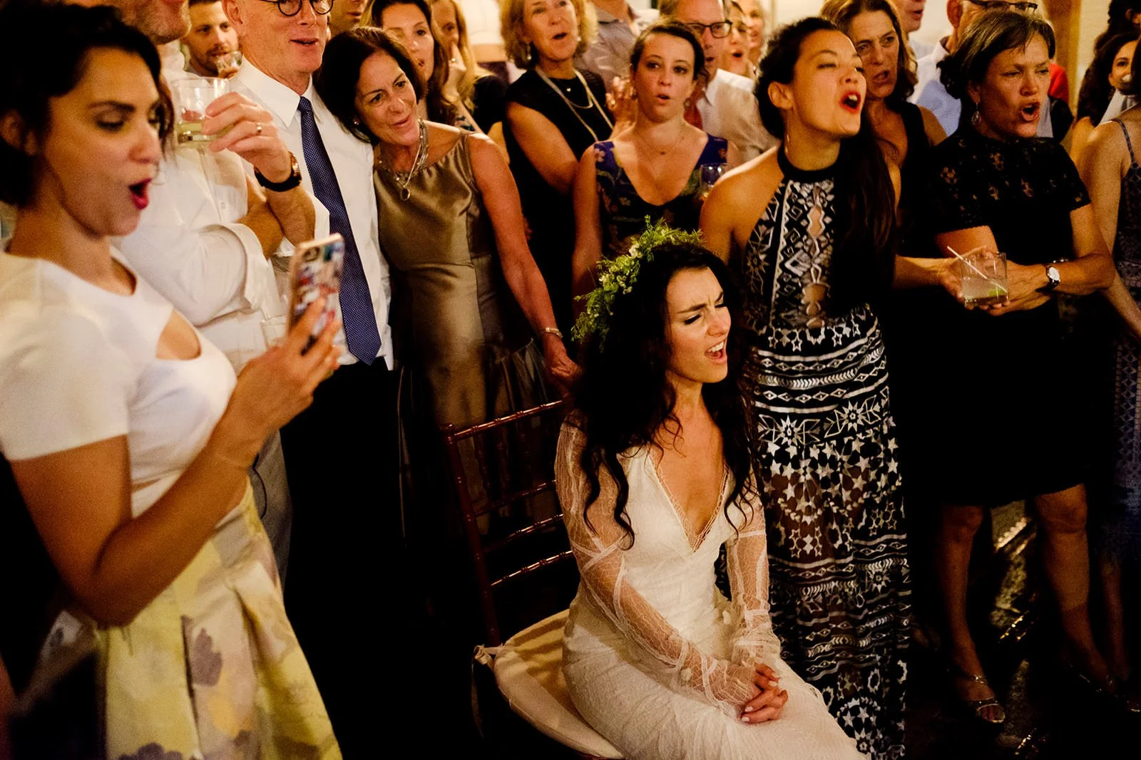 A wedding reception with a bride sitting, singing or crying, surrounded by guests standing and singing or singing along.