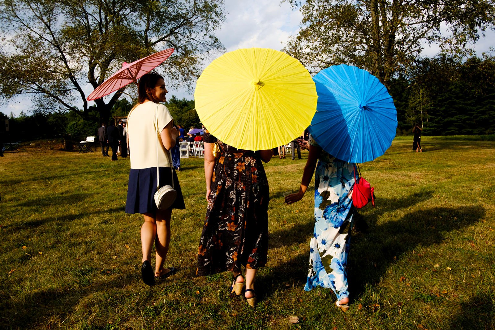 Three women walking on grass with two holding umbrellas, one pink, one yellow, one blue, at outdoor event on sunny day, with trees and gathering in background.