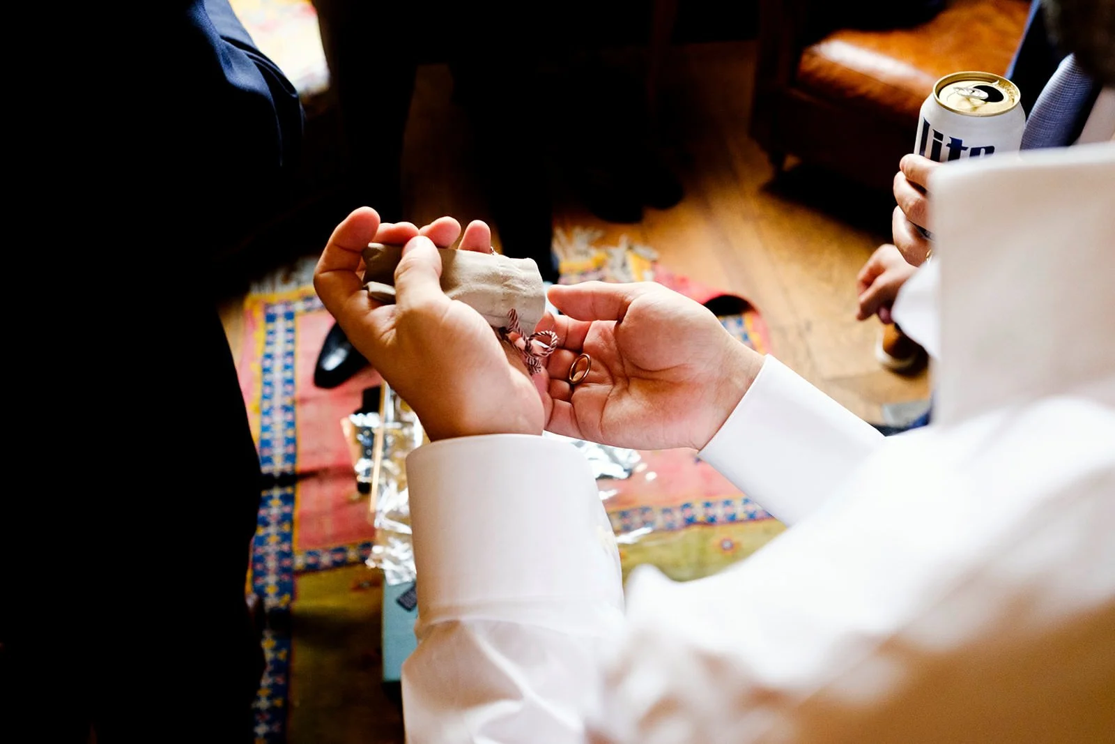 Close-up of a wedding ceremony where a person in a white shirt is placing a ring on a bride's finger during the exchange of vows. The bride is holding a small cloth pouch. A can of Miller Lite beer is visible in the background. The ceremony appears t