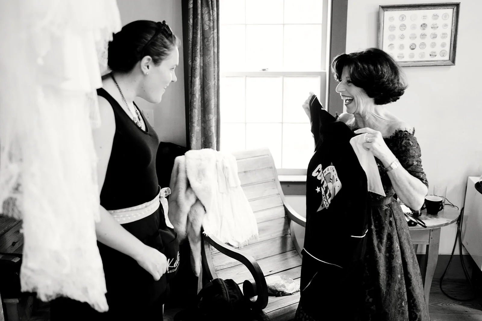 An elderly woman and a young woman are smiling and looking at each other; the elderly woman is holding up a dark jacket with a graphic print, in a well-lit room with a window, a wooden bench, and framed pictures on the wall.