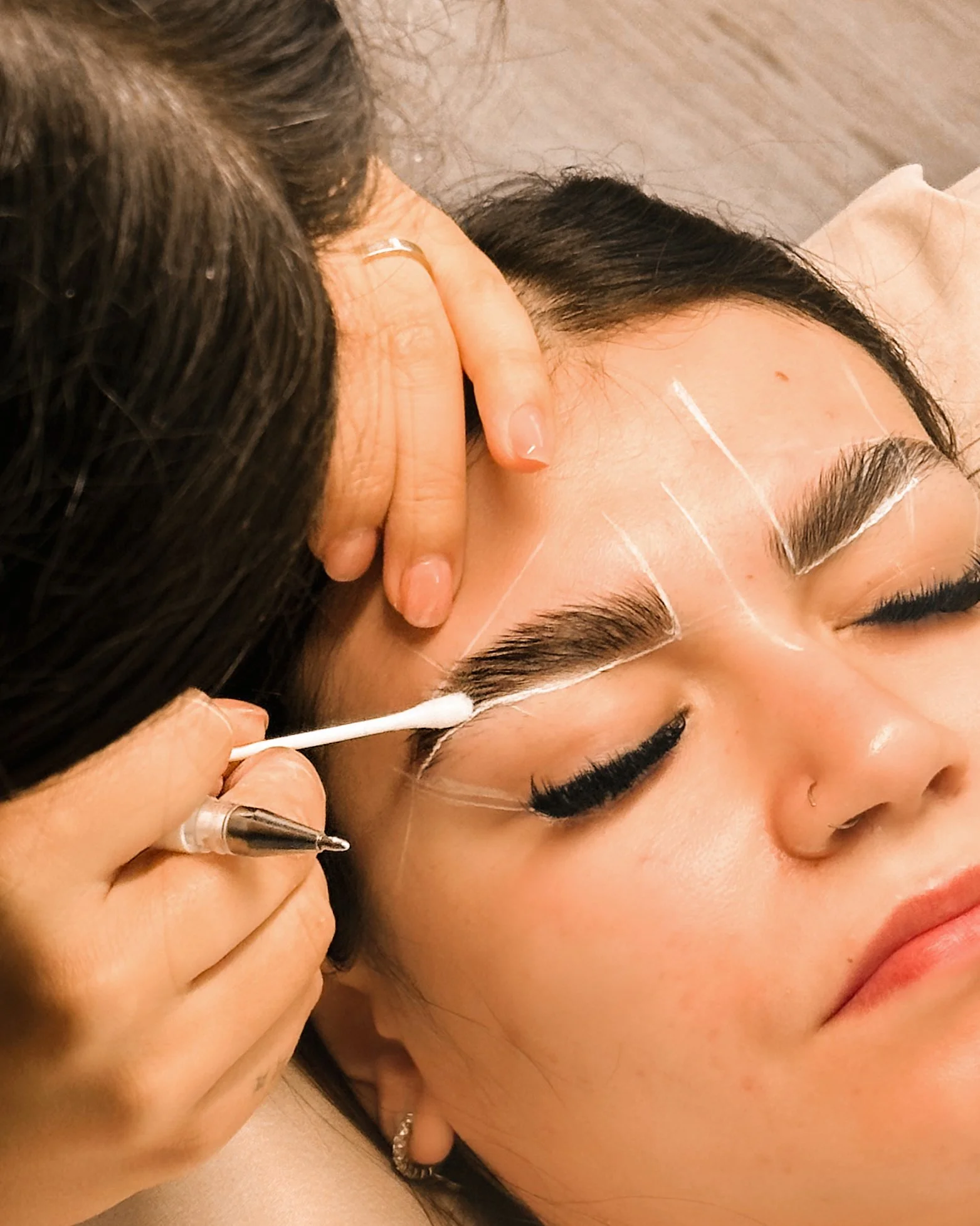 A woman receiving an eyelash or eyebrow treatment, lying down with her eyes closed as a technician applies makeup or semi-permanent pigment to her eyebrows.