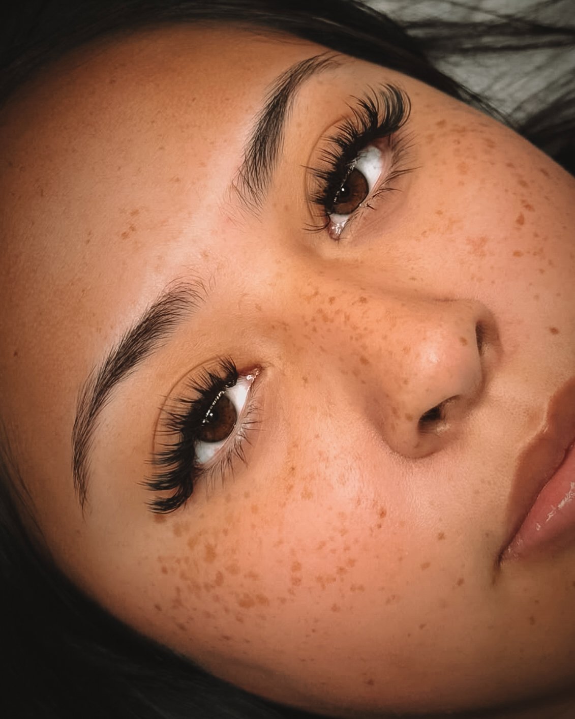 Close-up of a woman's face showing her brown eyes, long eyelashes, arched eyebrows, and numerous freckles on her cheeks and nose.