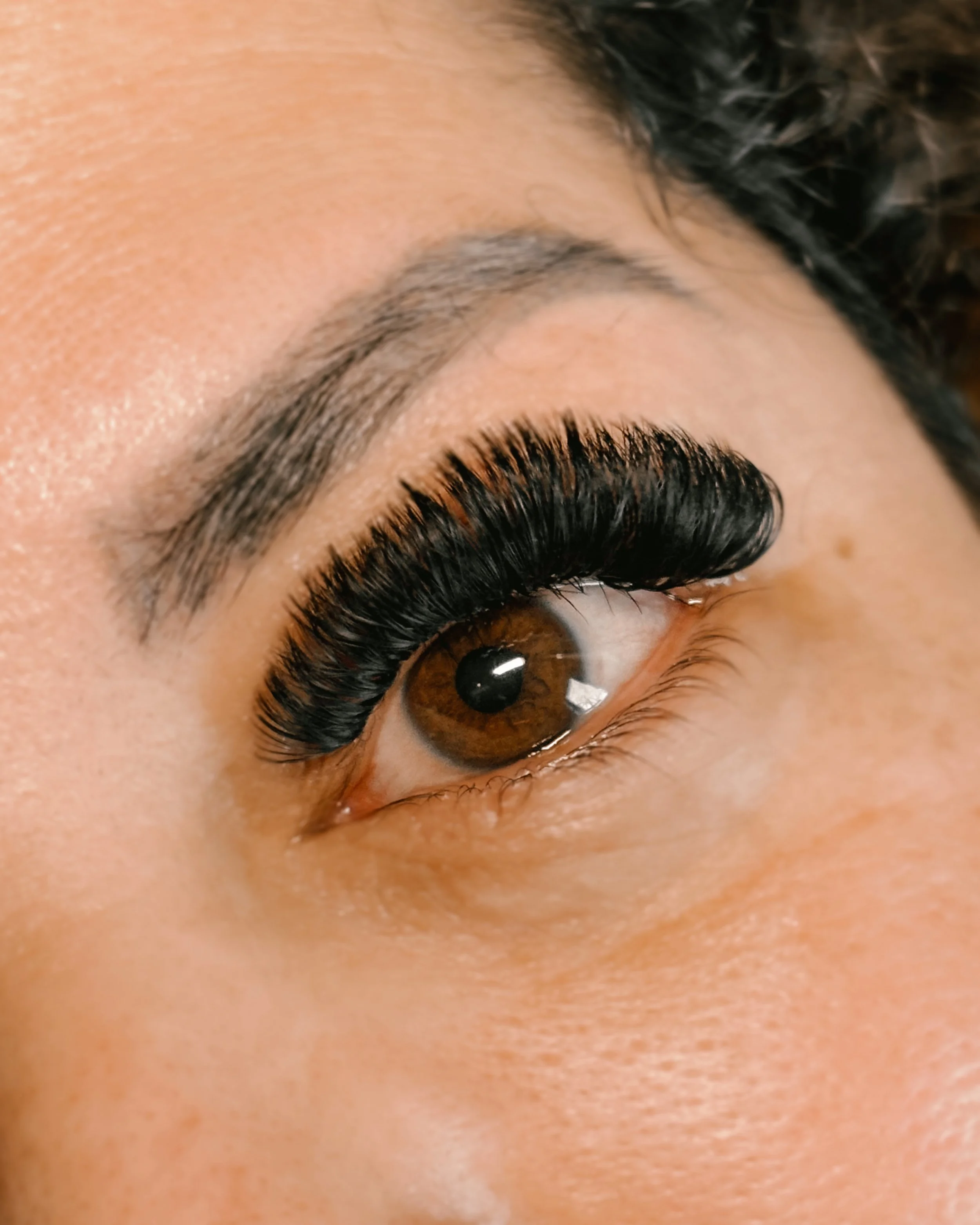 Close-up of a person's eye with long, thick, black eyelash extensions, brown iris, and well-groomed eyebrows.