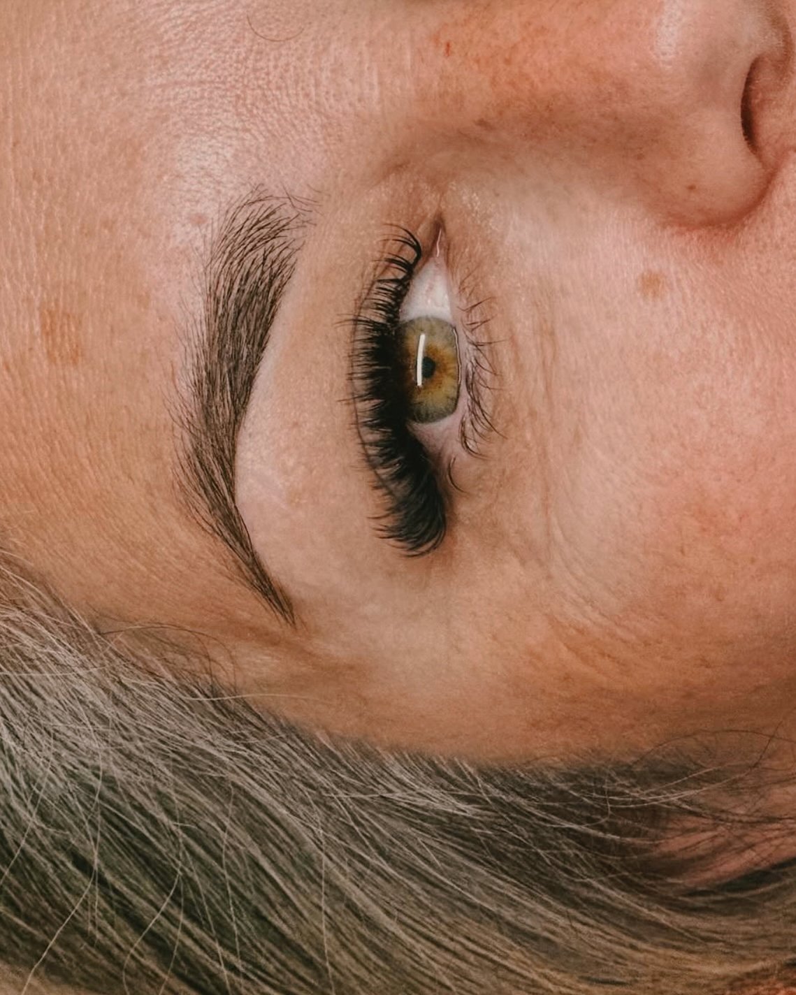 Close-up of a person with eye makeup, focusing on the eye with long, dark eyelashes and eyebrow, and a hint of blonde hair.