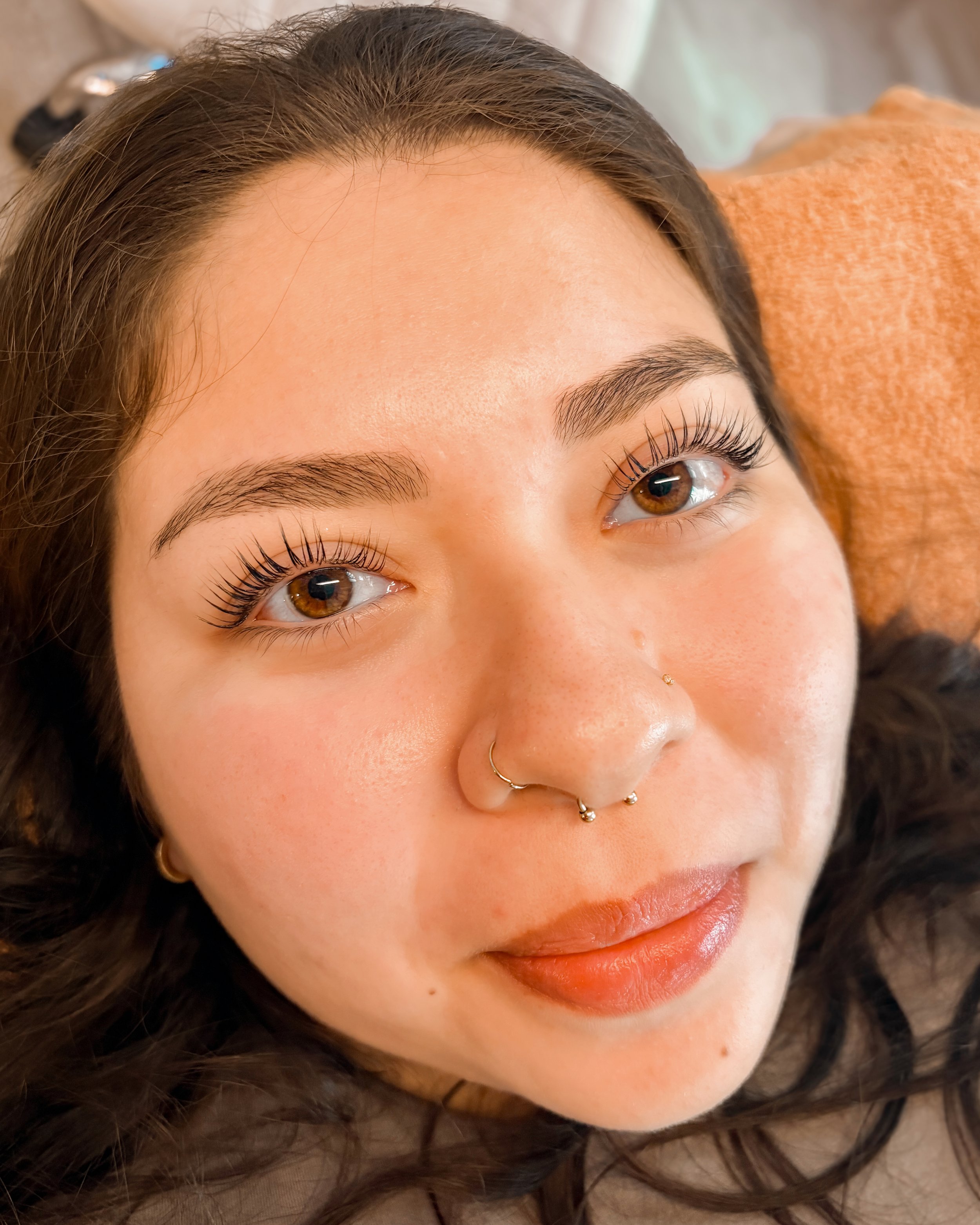 Close-up of a young woman with brown hair, brown eyes, and makeup. She has a nose piercing and septum jewelry, and is lying on an orange cushion.