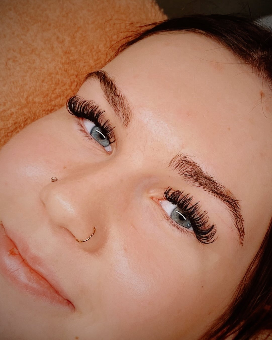 Close-up of a woman's face with eye makeup, including long, thick false eyelashes, well-defined eyebrows, and a nose piercing.