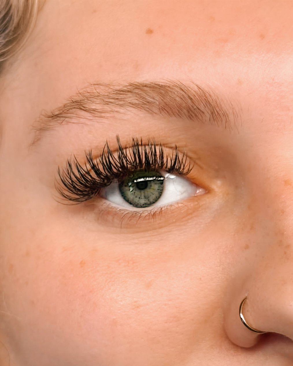 Close-up of a person's eye with long, thick eyelashes, a well-groomed eyebrow, and a gold nose ring.