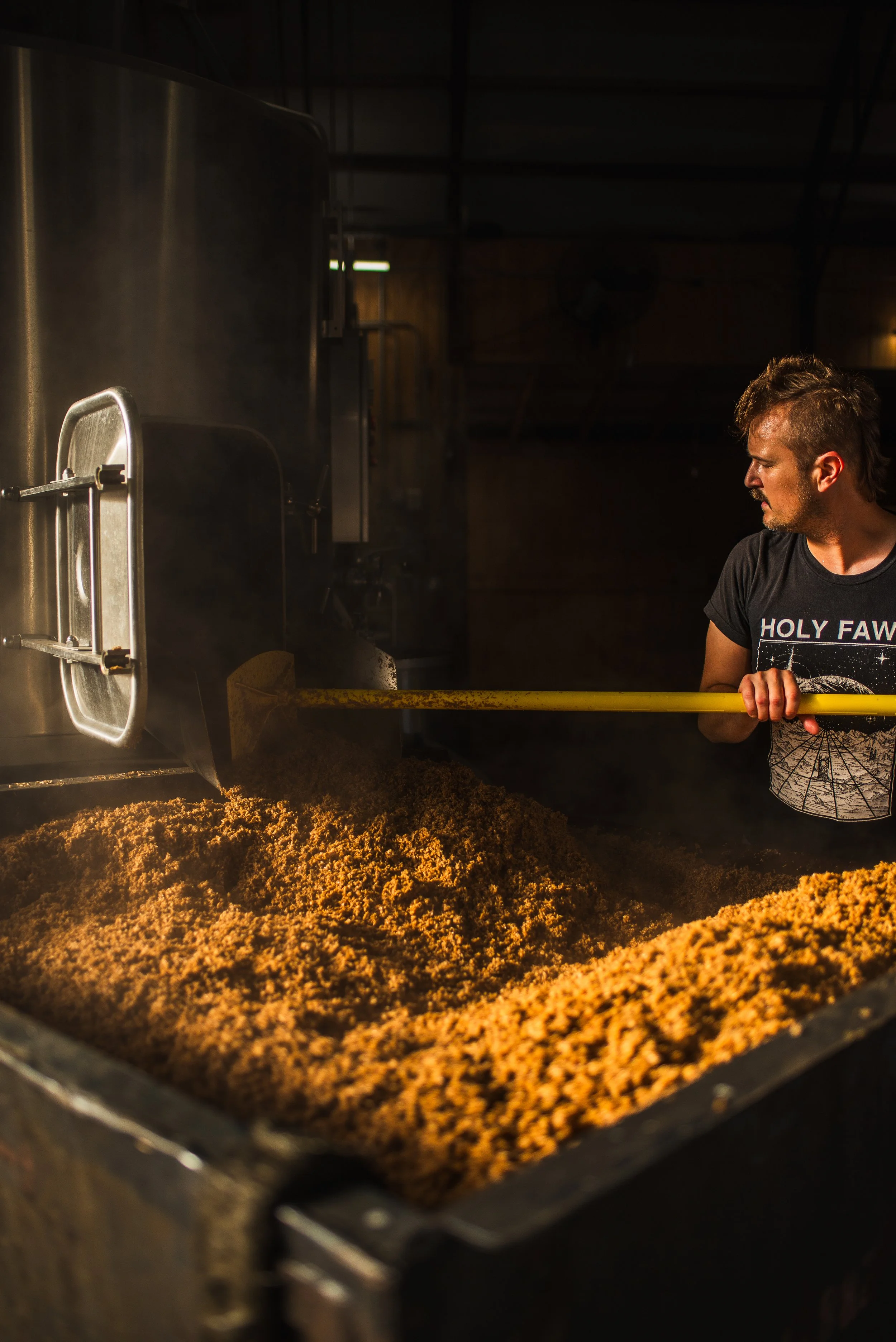 A man with a beard and wearing a black t-shirt with the words 'HOLY FAWKS' stands next to a large metal container filled with a brown, crumbly material, in a dimly lit industrial environment.
