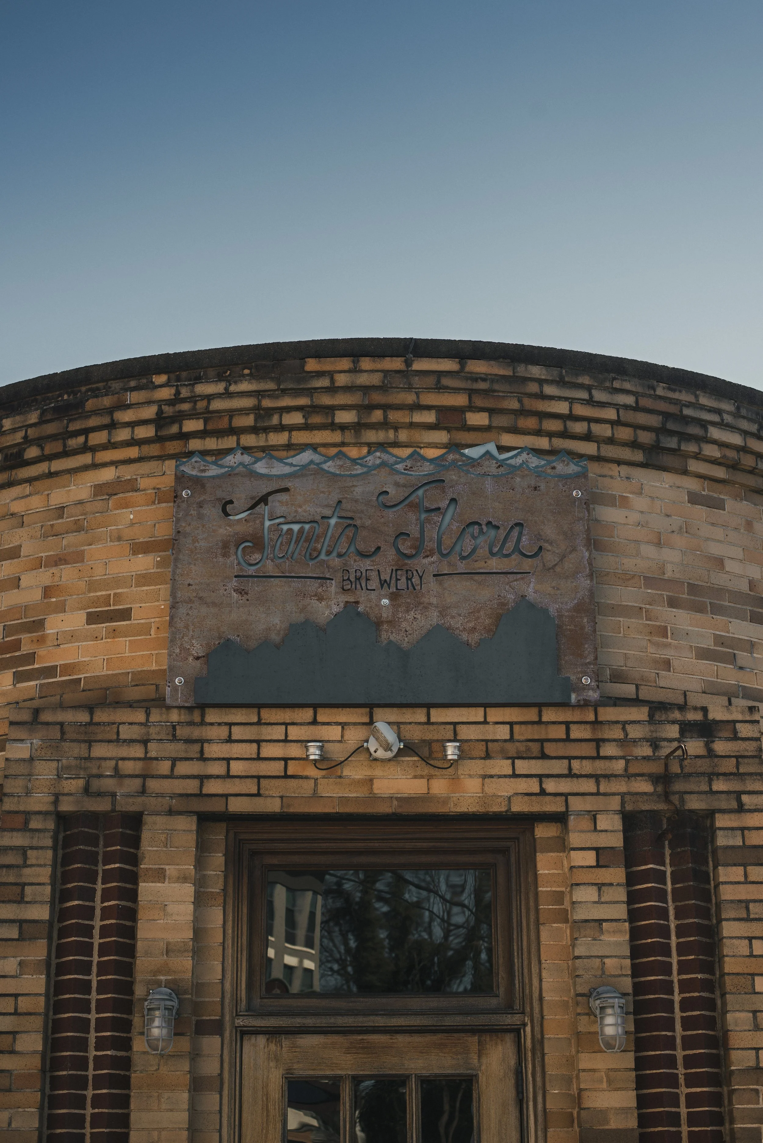 Close-up of a sign on a brick building that reads 'Fonta Flora Brewery' with decorative waves above the text, and a window below, with trees and sky reflected in the window.