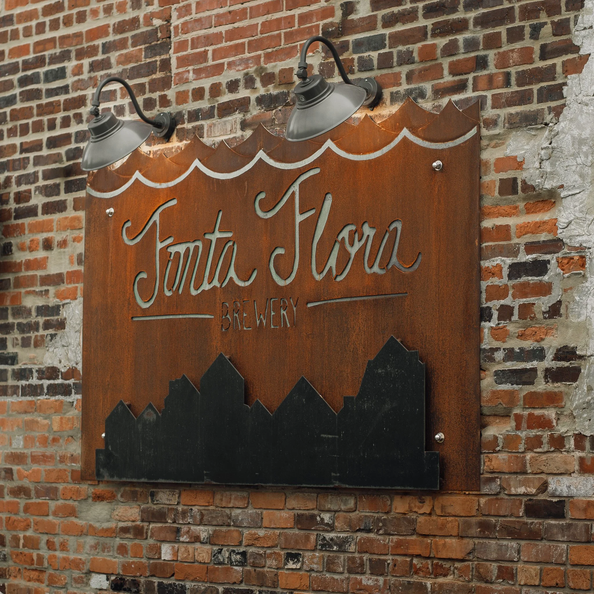 Close-up of a brick wall with a rustic metal sign that reads 'Fonta Flora' illuminated by two industrial-style wall-mounted lights.