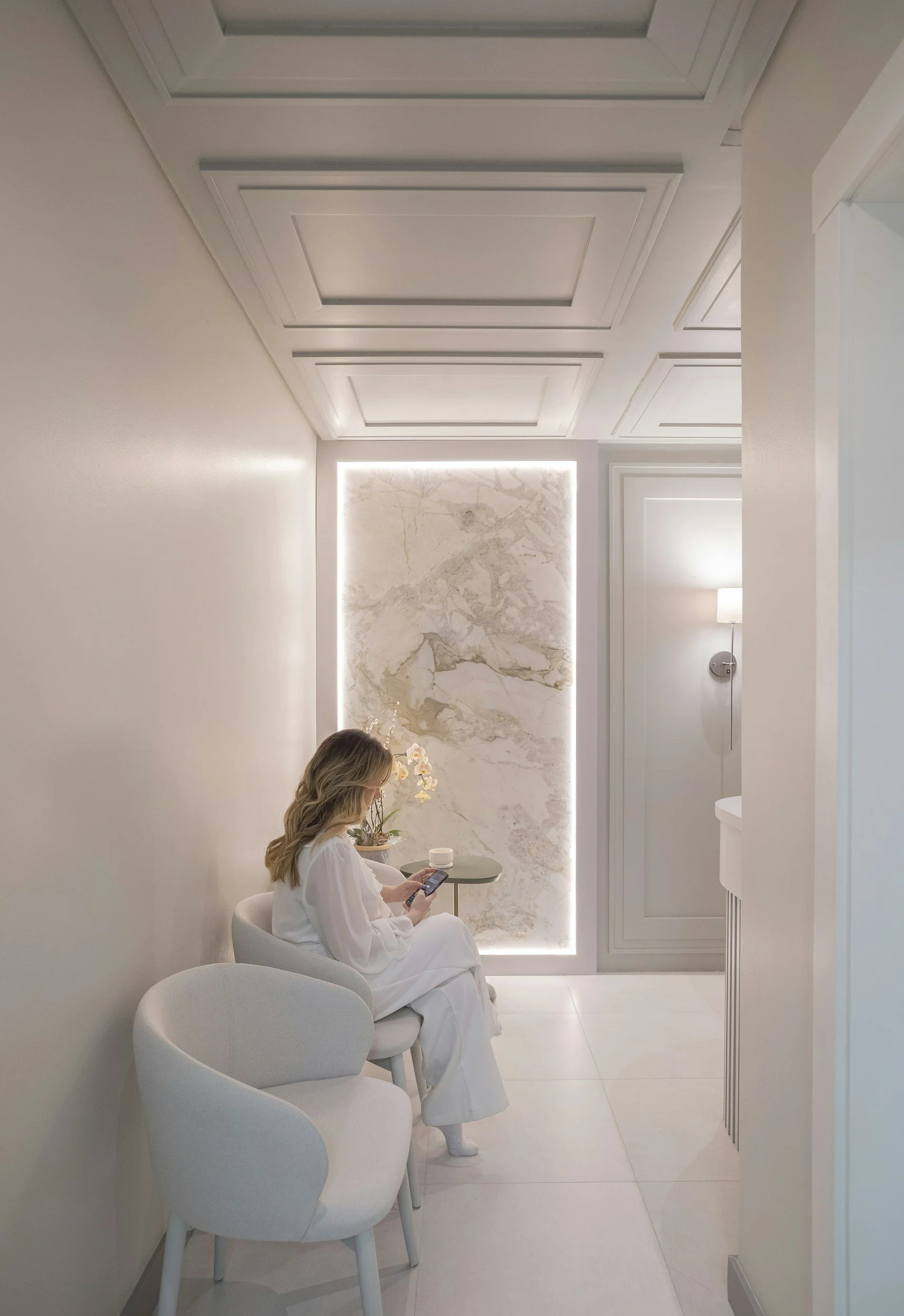 A woman in white clothing sitting in a modern, minimalist room on a light-colored chair, looking at her phone, with a small side table holding flowers and a candle in front of a backlit marble wall panel.