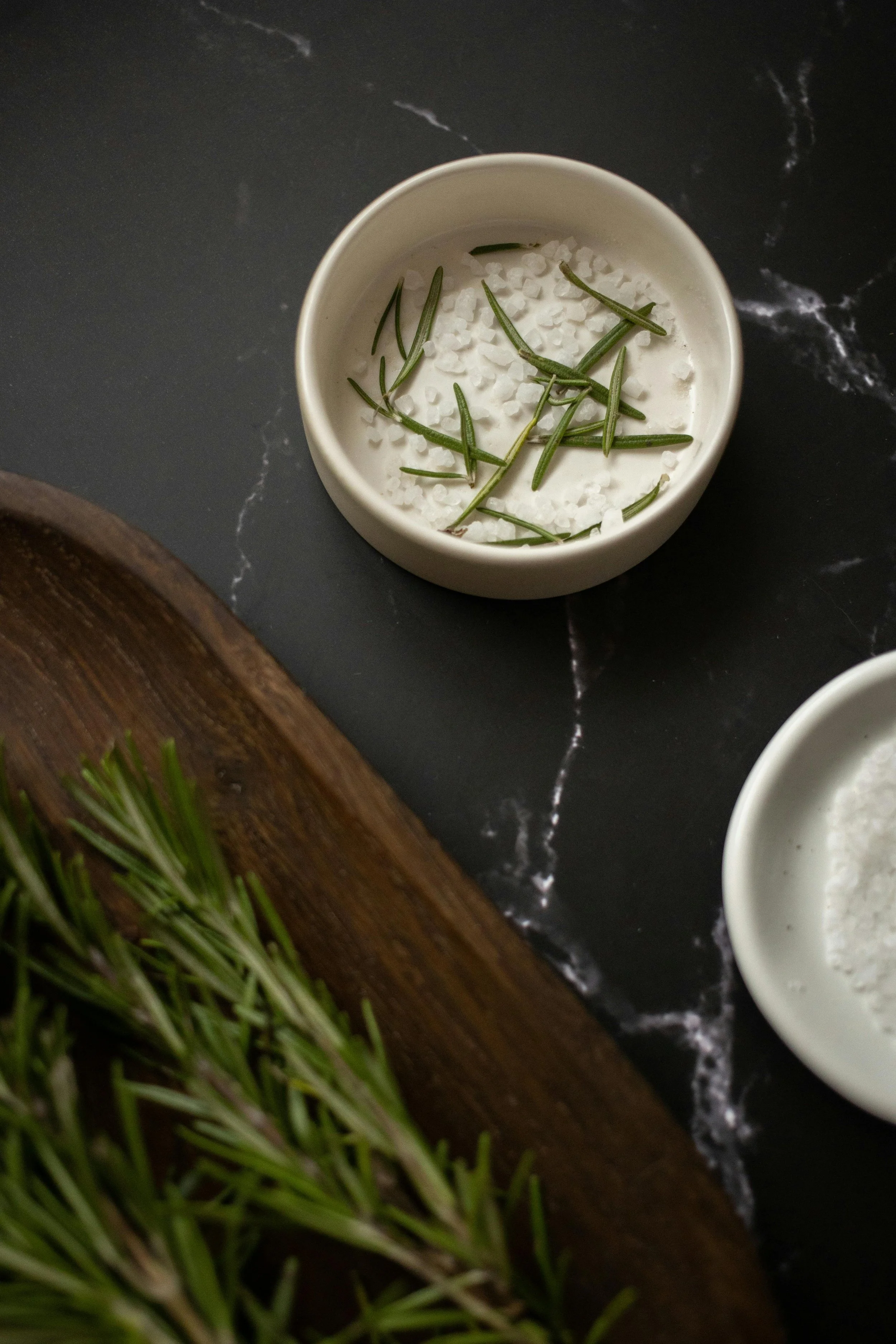 White bowl containing coarse sea salt and sprigs of fresh rosemary on a dark marble surface, with part of a wooden board and additional rosemary in the corner.