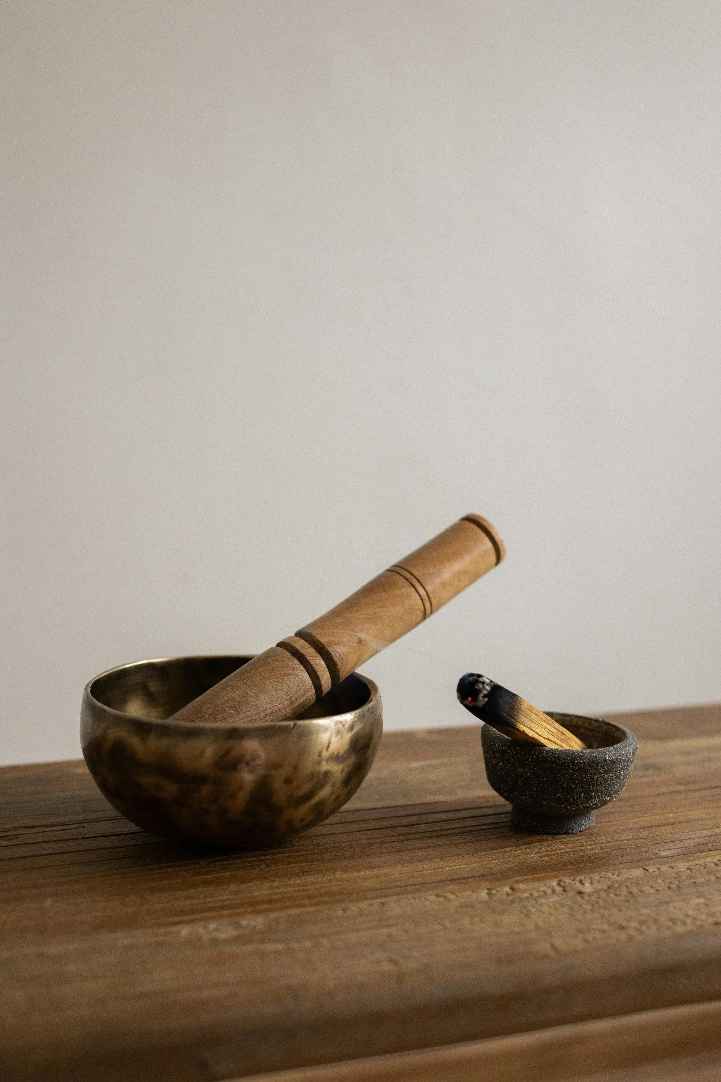 Brass singing bowl with a wooden mallet resting inside, and a smaller dark stone bowl with a lit stick of incense, placed on a wooden surface.