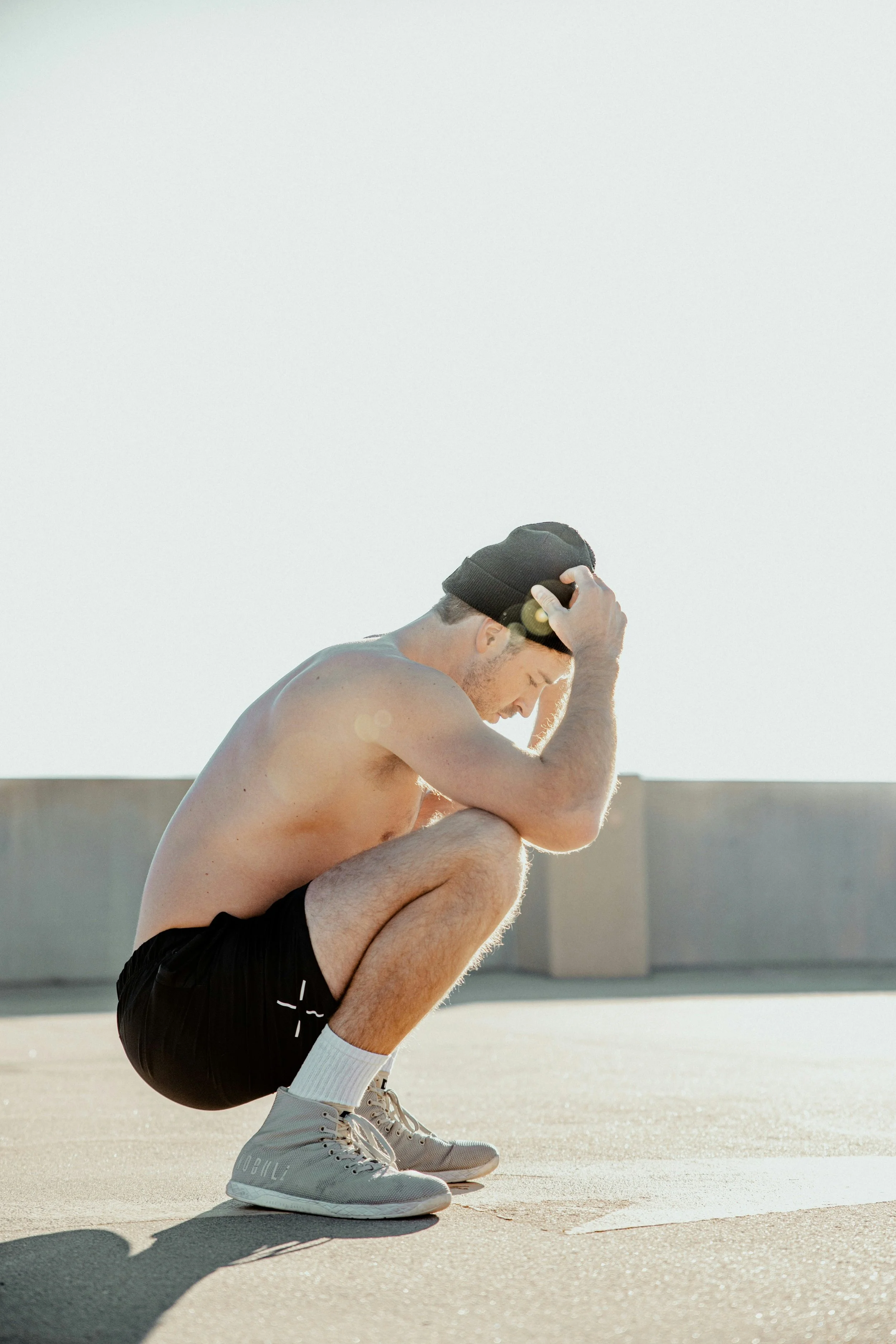 A shirtless man wearing black shorts, white socks, and gray sneakers crouches on a concrete surface with his head bowed and hands on his head, outdoors under bright sunlight.
