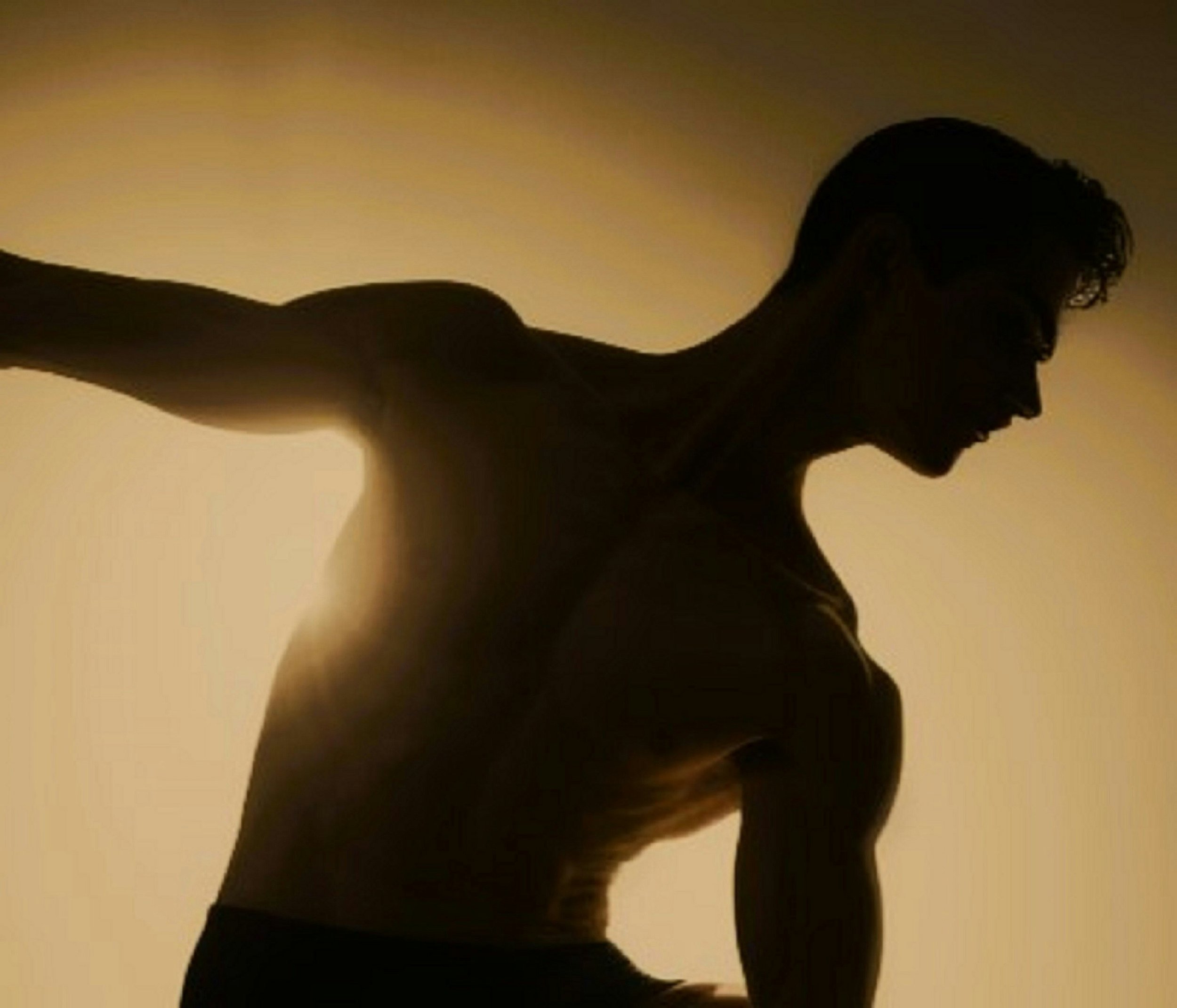 Silhouette of a shirtless man with muscular arms and curly hair, facing downward, backlit with a warm light.