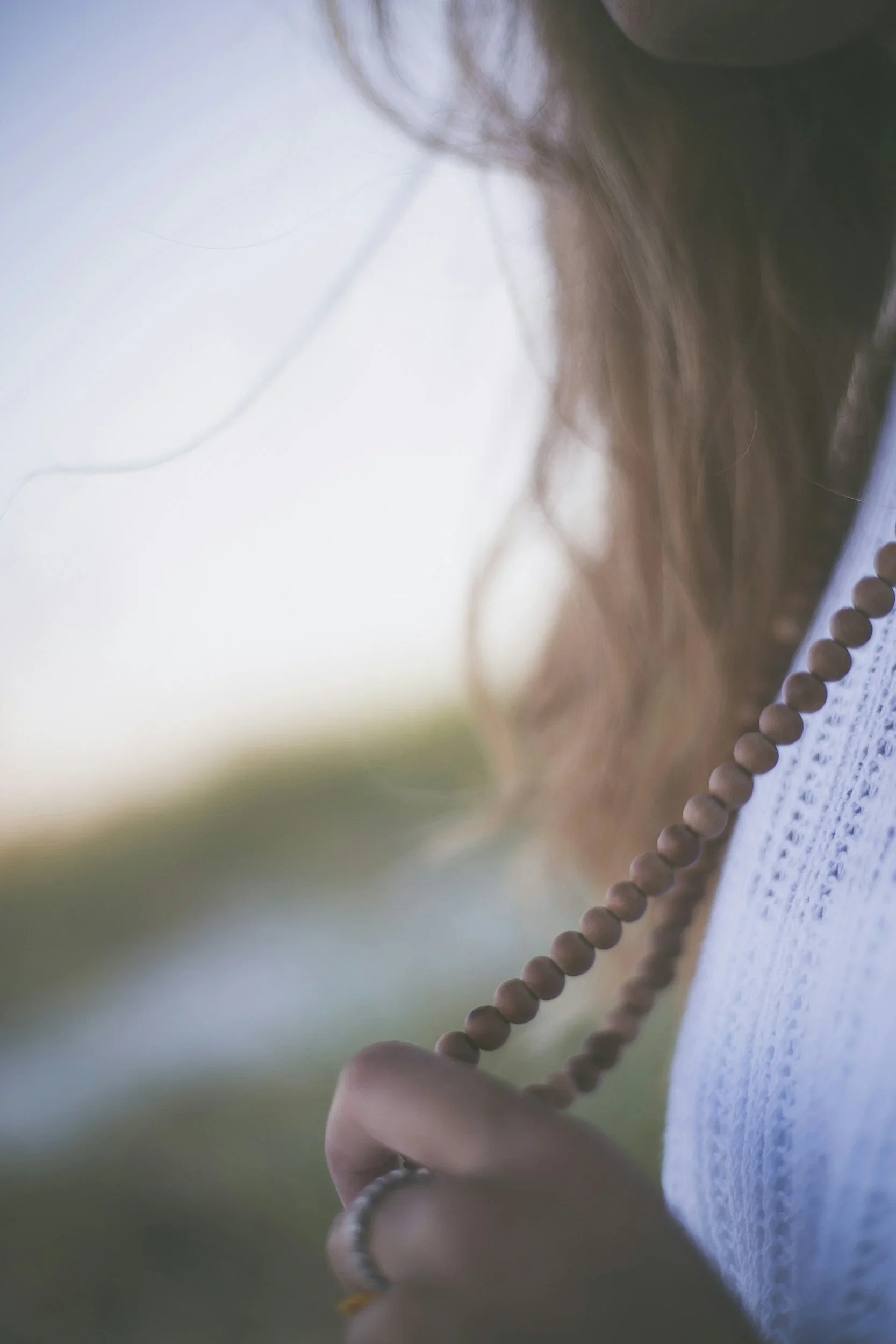 A person holding a wooden beaded necklace near their chin, with a blurred outdoor background.