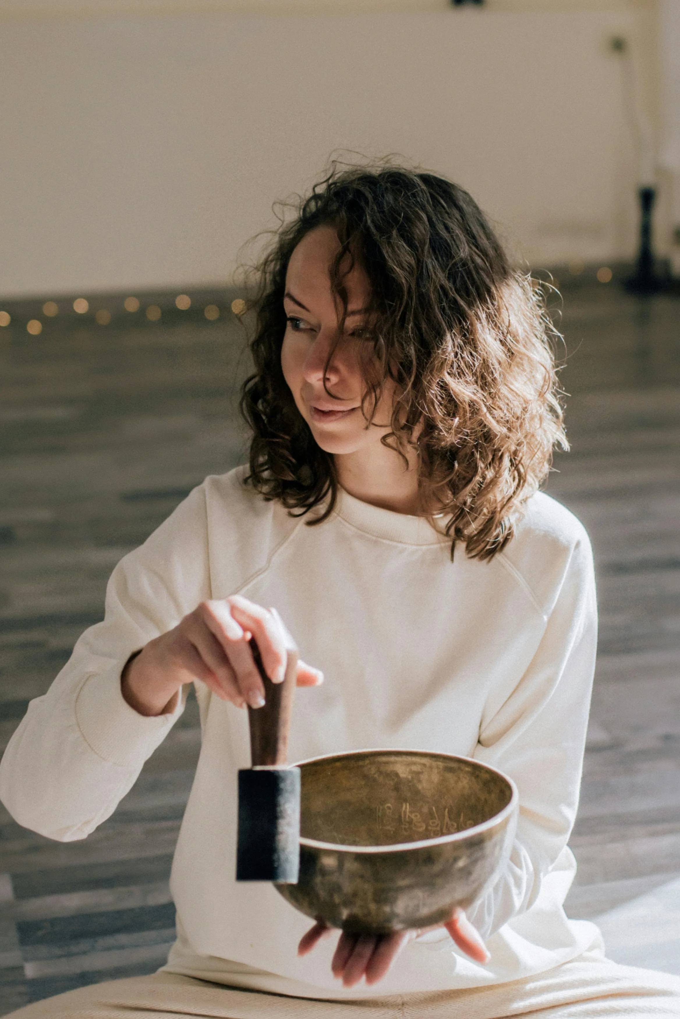 A woman with curly hair in a cream sweatshirt sitting on a wooden floor, holding a singing bowl with a mallet, looking to her left.