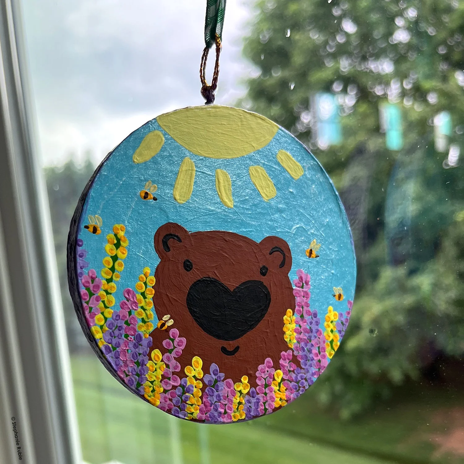 round ornament with green and white ribbon hanging in window. painted image on ornament is a bear face peeking out from a field of flowers, with a big sun overhead.