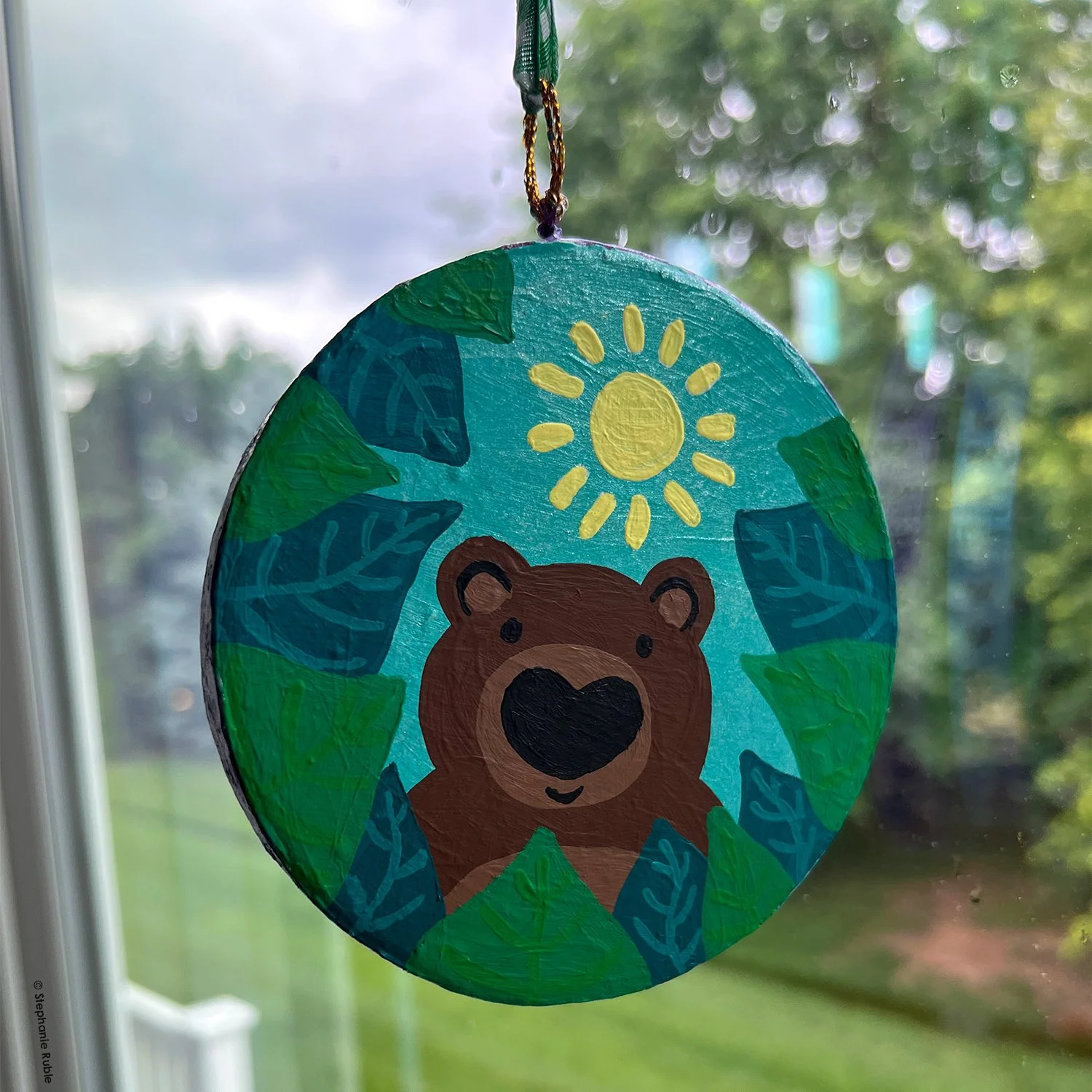 round ornament with green and white ribbon hanging in window. painted image on ornament is a bear looking out from a sea of green leaves, with a small sun above them.