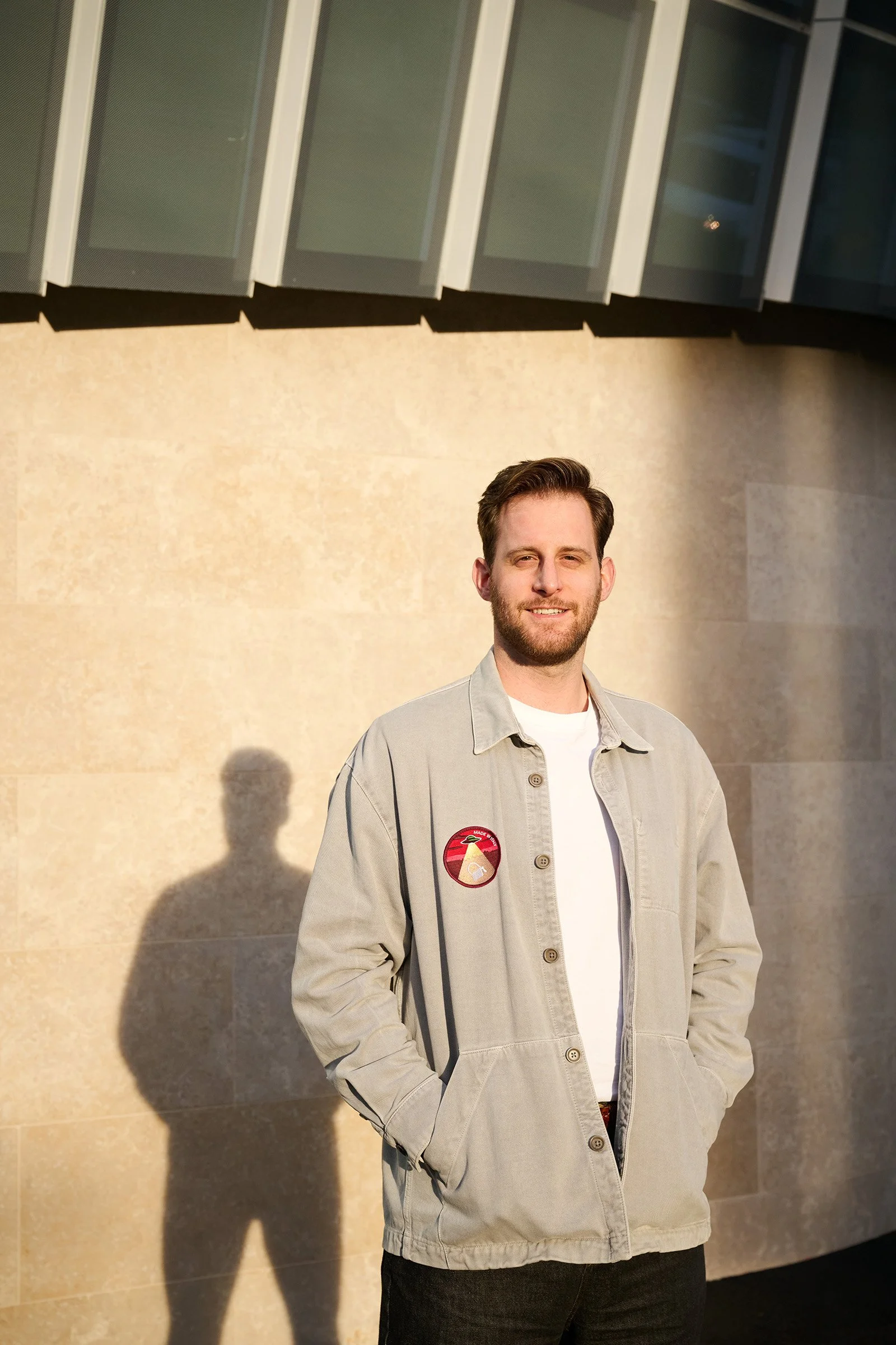 A young man with a beard and short hair standing outdoors against a beige brick wall, wearing a light jacket and smiling at the camera, with his hands in his pockets and a shadow of himself cast on the wall.