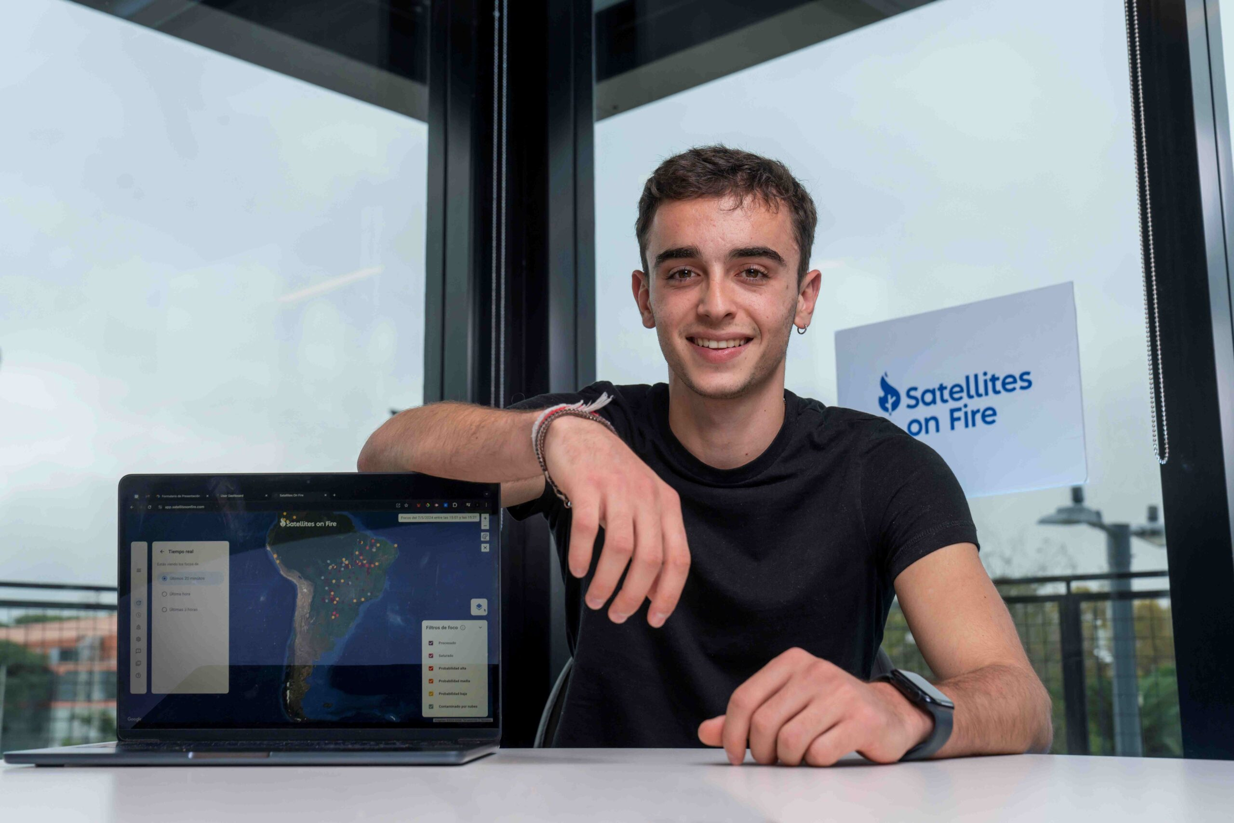A young man sitting at a table, smiling, with a laptop showing a map of South America. There is a sign that reads 'Satellites on Fire' behind him.