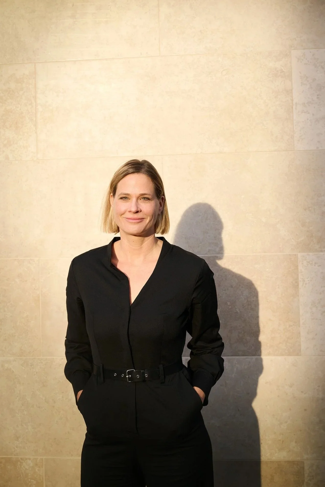 A woman with short blonde hair wearing a black outfit and standing against a beige tiled wall, casting a shadow.