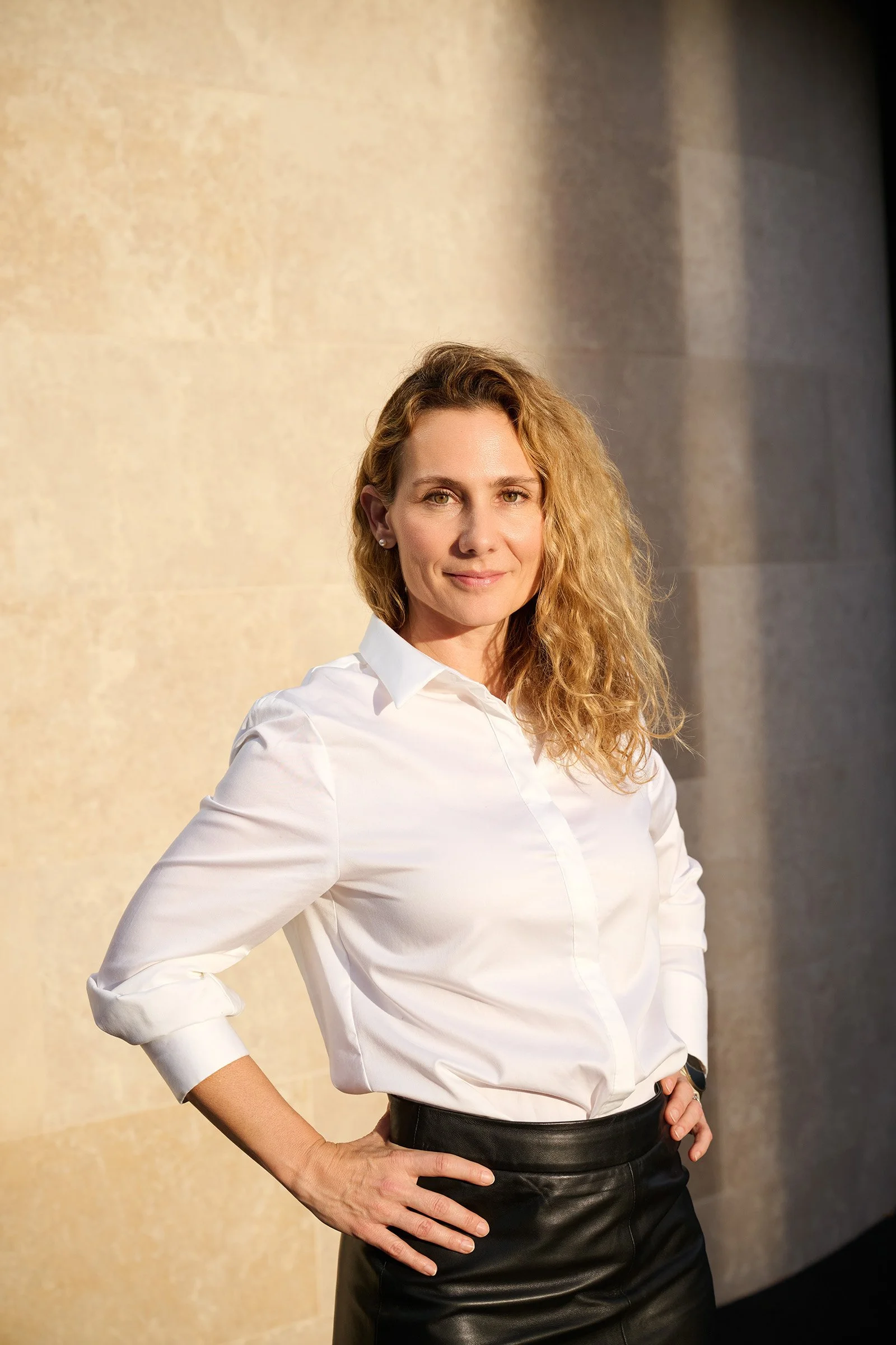 A woman with wavy blonde hair poses confidently against a beige stone wall, wearing a white button-up shirt and black leather skirt.