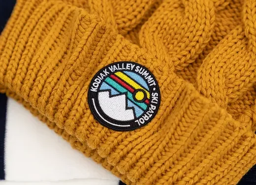 Close-up of a mustard yellow knitted hat with a round patch that reads 'Kobuk Valley Summit 2023' and features colorful geometric design.