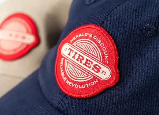 Close-up of a navy blue cap with a red and white circular patch that says "Herald's County Tires" and "Dubble Revolution." In the background, there is a blurred patch with similar design.