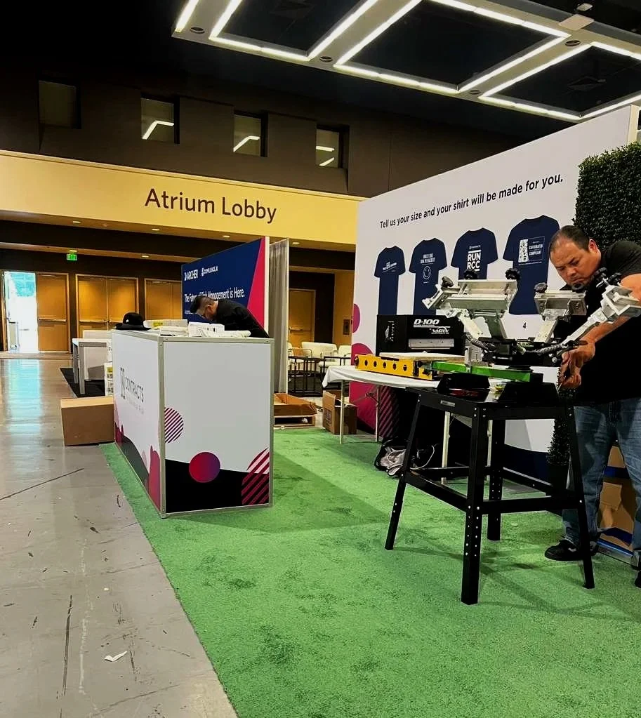 Silent auction booth set up in a convention center, with a man adjusting a mechanical device on a table, and a large banner showing t-shirt size and design. The booth is on a green carpet, with a sign pointing to the Atrium Lobby in the background.