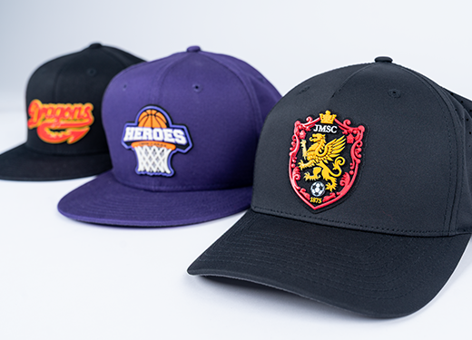 Three baseball caps with different team logos displayed in a row on a white background. The first cap is black with a yellow and red logo, the second is purple with a basketball and net logo, and the third is black with a red and gold lion emblem.