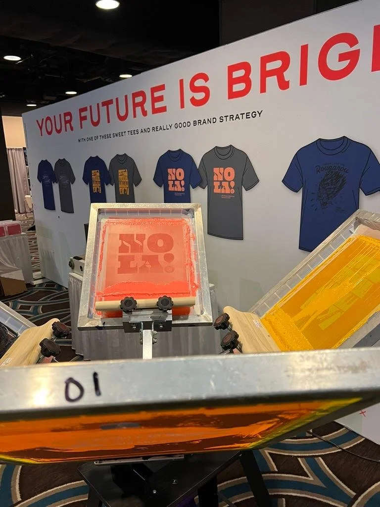 Screen printing machine with a pink print in progress, displaying the words "NO FEAR" and yellow ink roller to the side, at a booth with a banner that says "Your future is bright" and showcases various t-shirts.