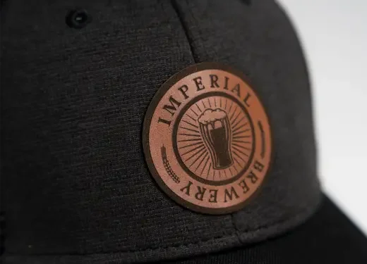 Close-up of a black hat with a round leather patch featuring a beer mug graphic and the words 'Imperial Brewery'.