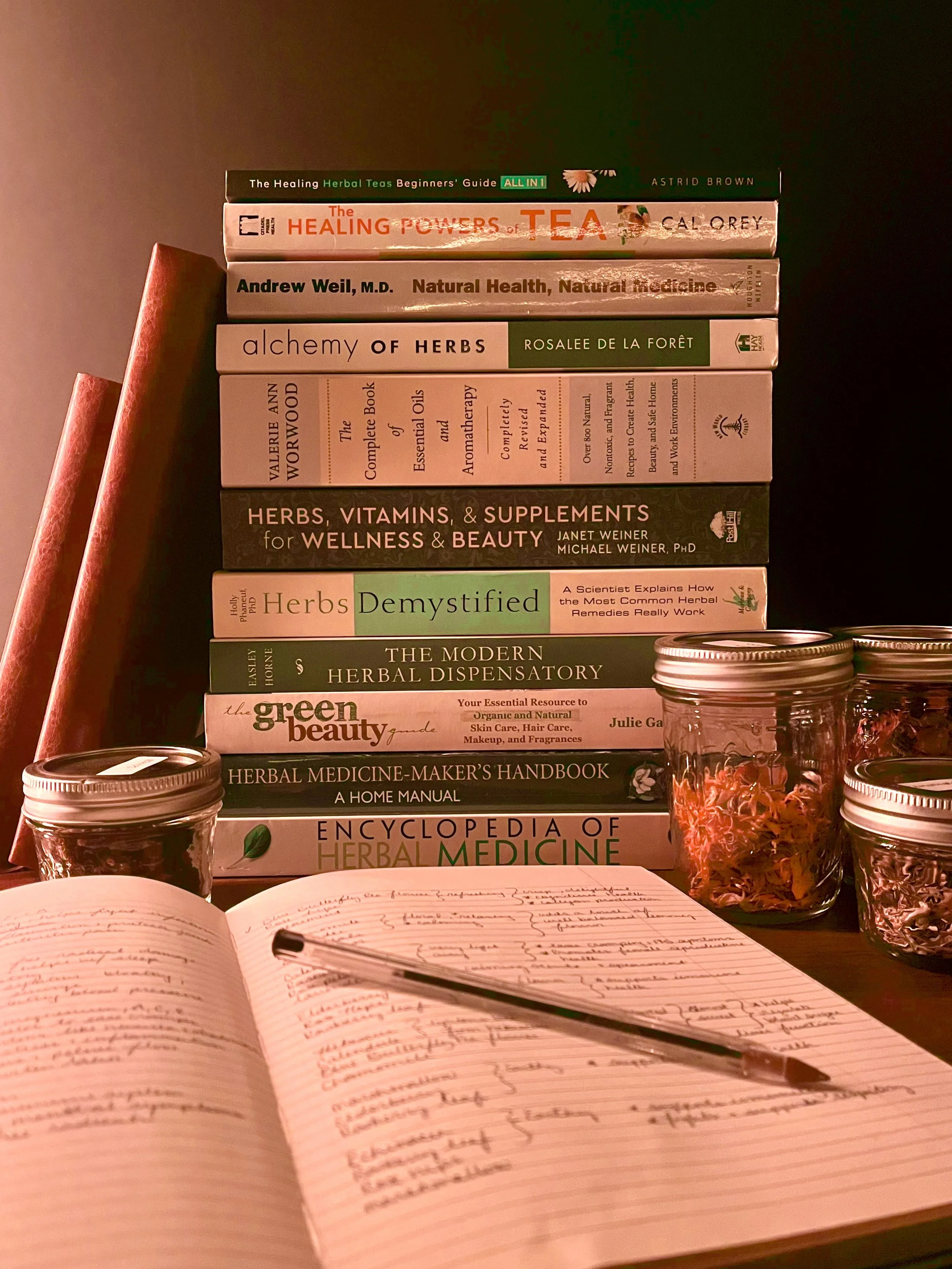 Stack of herbal medicine and health books on a desk with an open notebook and a pen, jars of herbs in front.
