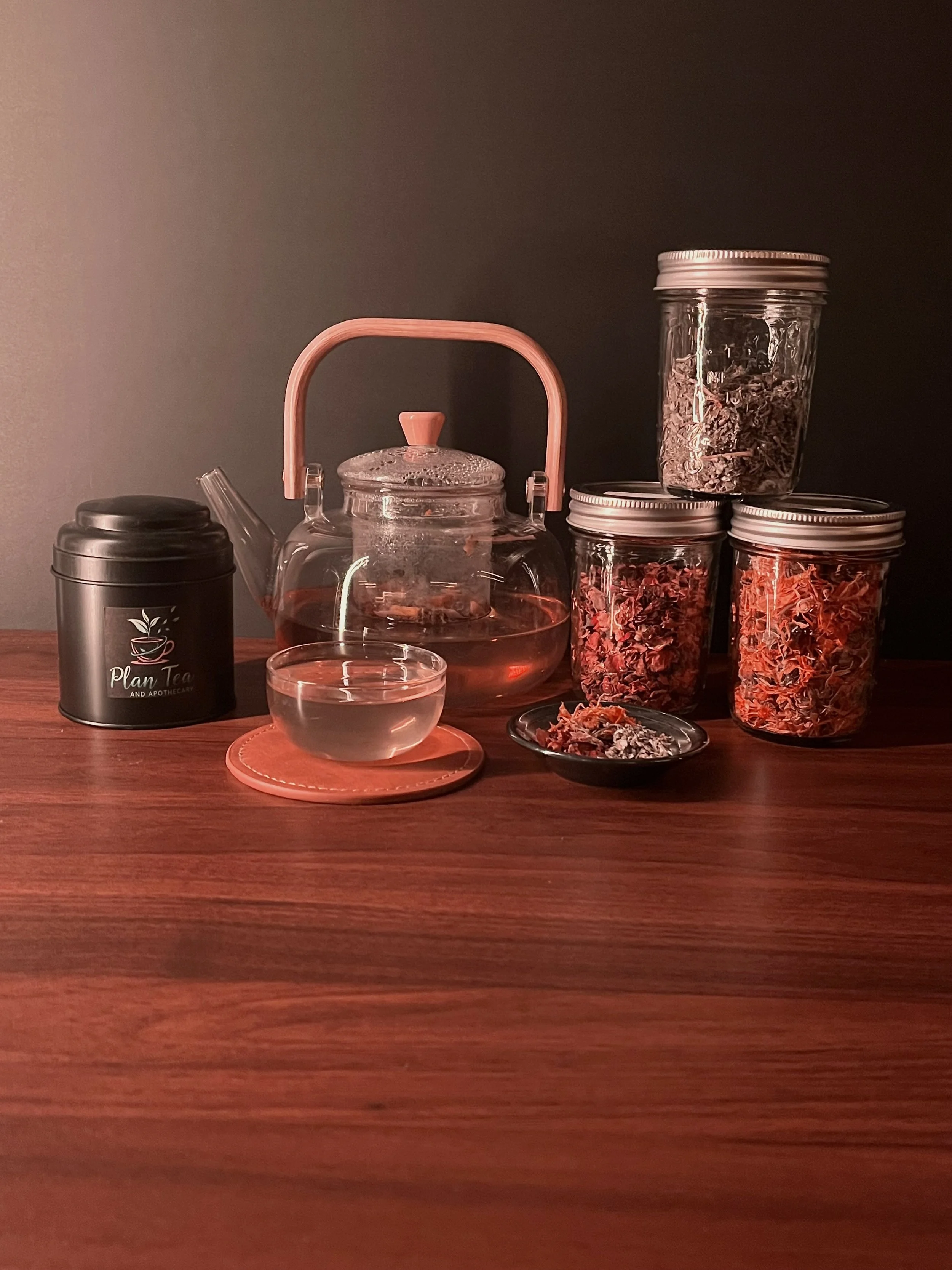 A wooden table holding a black tin can with a tea leaf logo, a transparent glass teapot with a pink handle and lid, a small glass teacup, and several glass jars filled with dried tea leaves or herbs. The background is dark and plain.