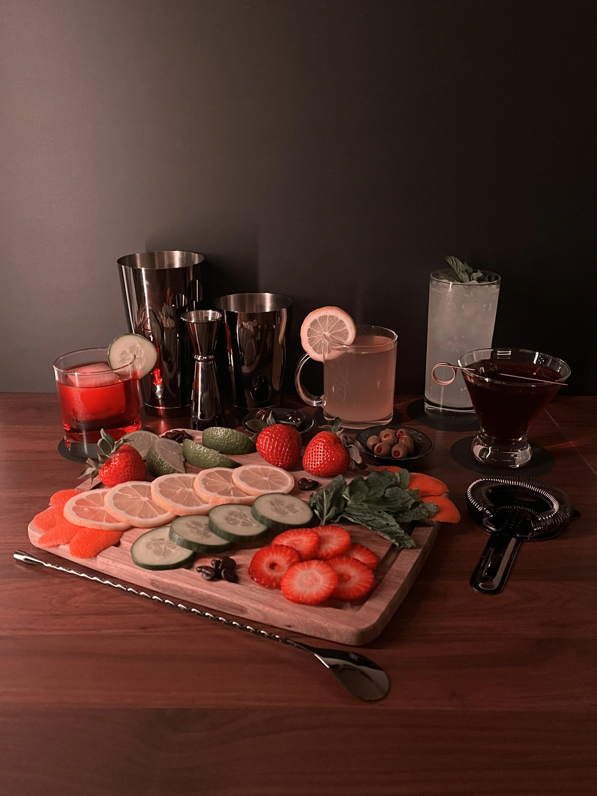 A wooden board with sliced strawberries, cucumbers, lemon slices, and strawberries on it, surrounded by various cocktails and bar accessories on a dark wooden table against a black background.