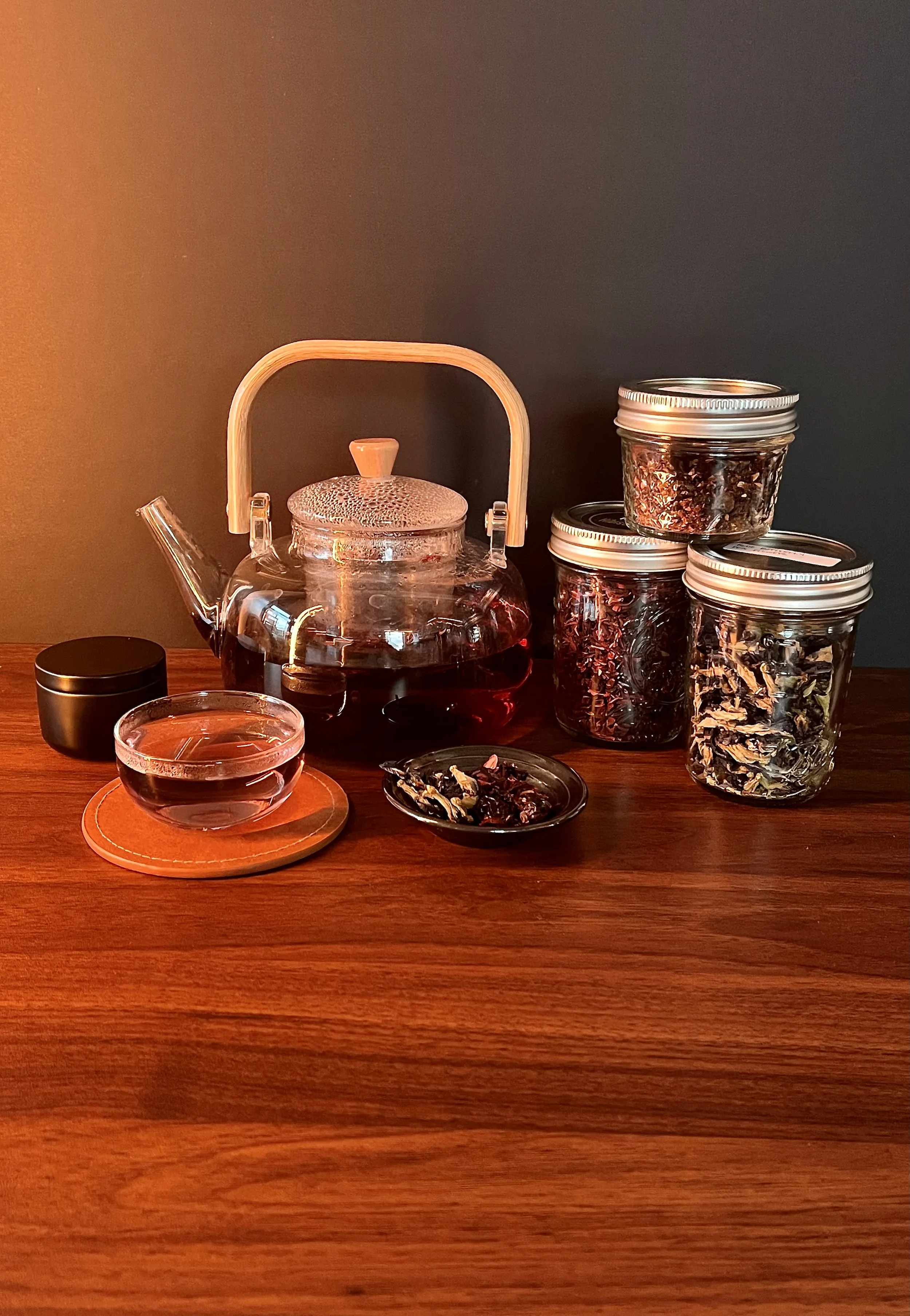 A glass teapot filled with tea, surrounded by jars of dried herbs and spices, a small bowl of dried tea leaves, and a black container on a wooden surface against a dark background.
