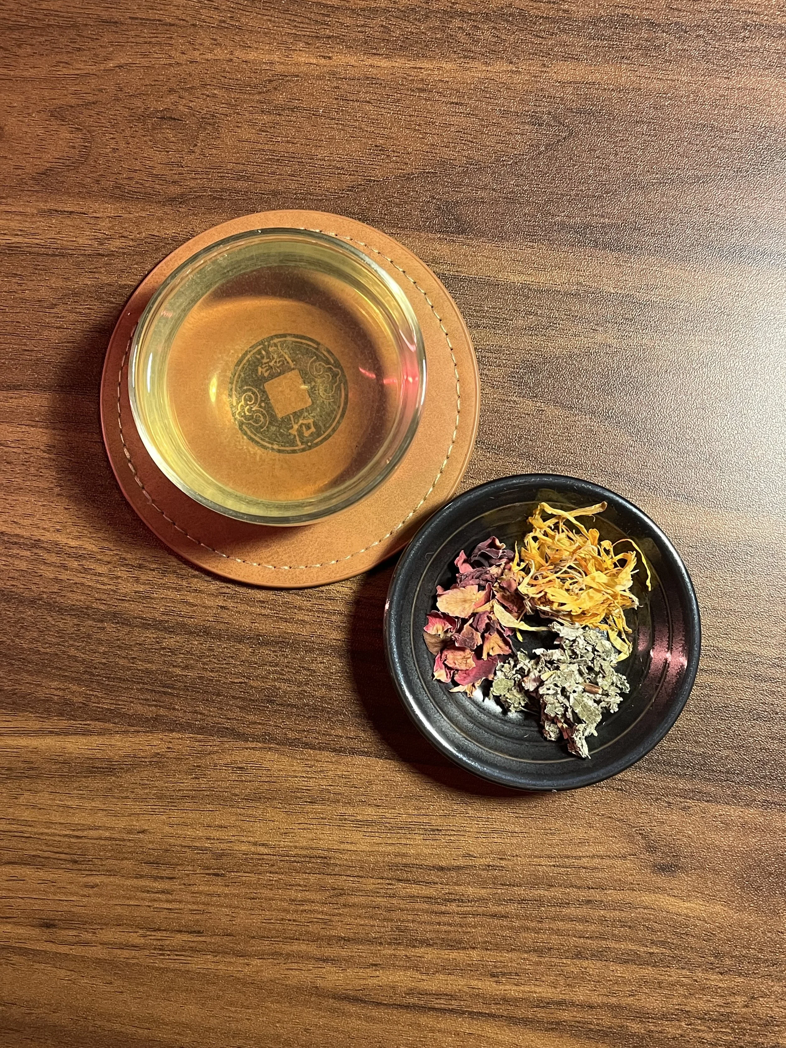 A glass of tea on a leather coaster and a black dish with dried flowers on a wooden table.